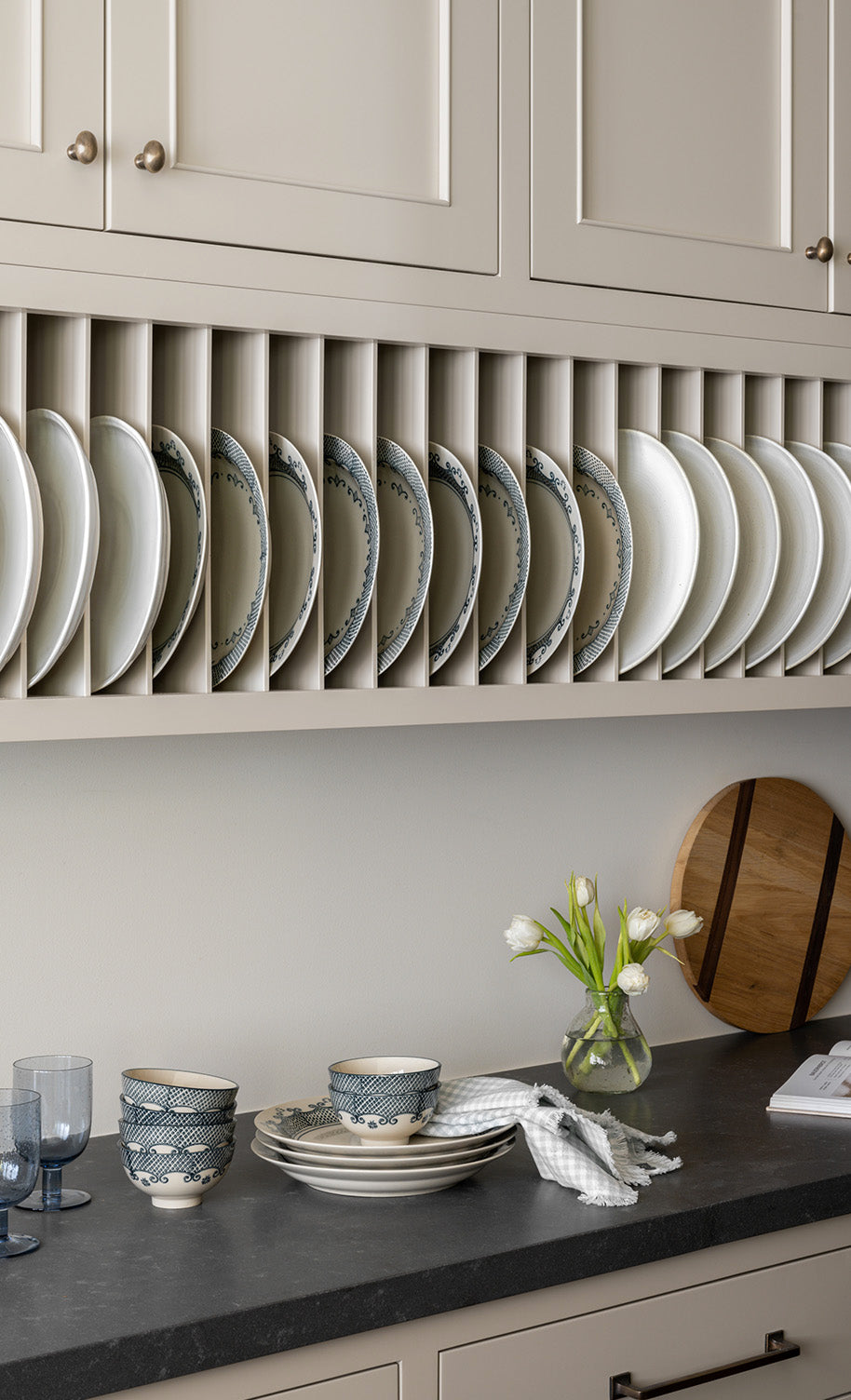 Plates are neatly arranged vertically in a rack under beige kitchen cabinets. A countertop below holds bowls, stacked plates, two blue glasses, a vase with white tulips, and a wooden cutting board, by McGee & Co.