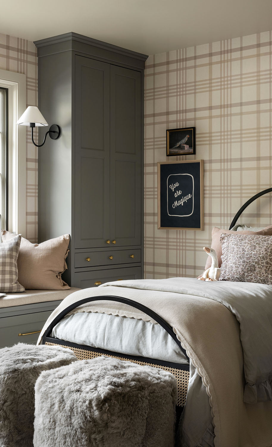 A cozy bedroom with plaid wallpaper, a bed with beige and gray bedding, a window seat with pillows, a gray wardrobe, wall art, a wall lamp, and two furry stools at the foot of the bed, by McGee & Co.