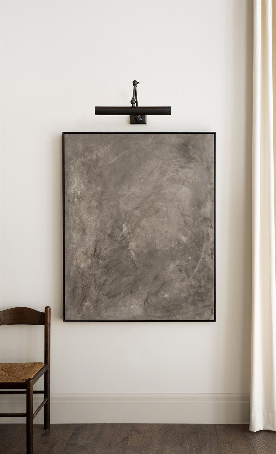 A large, abstract gray painting with subtle texture hangs on a white wall under a black picture light. A wooden chair with a woven seat is partially visible to the left, and a cream curtain is to the right, by McGee & Co.