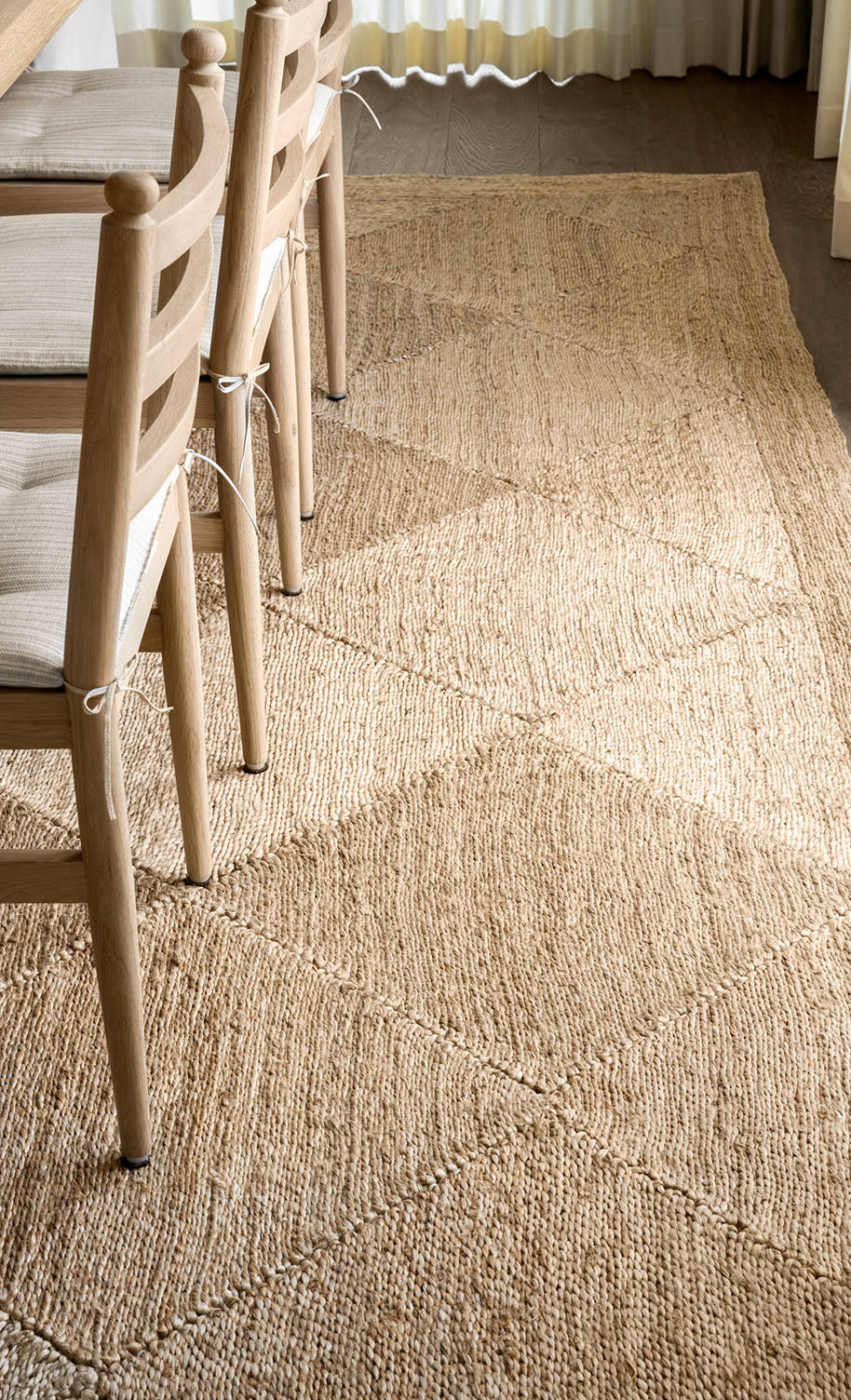 A close-up view of a textured, woven beige rug beneath wooden chairs with light-colored seat cushions, placed next to a table in a sunlit room by McGee & Co.