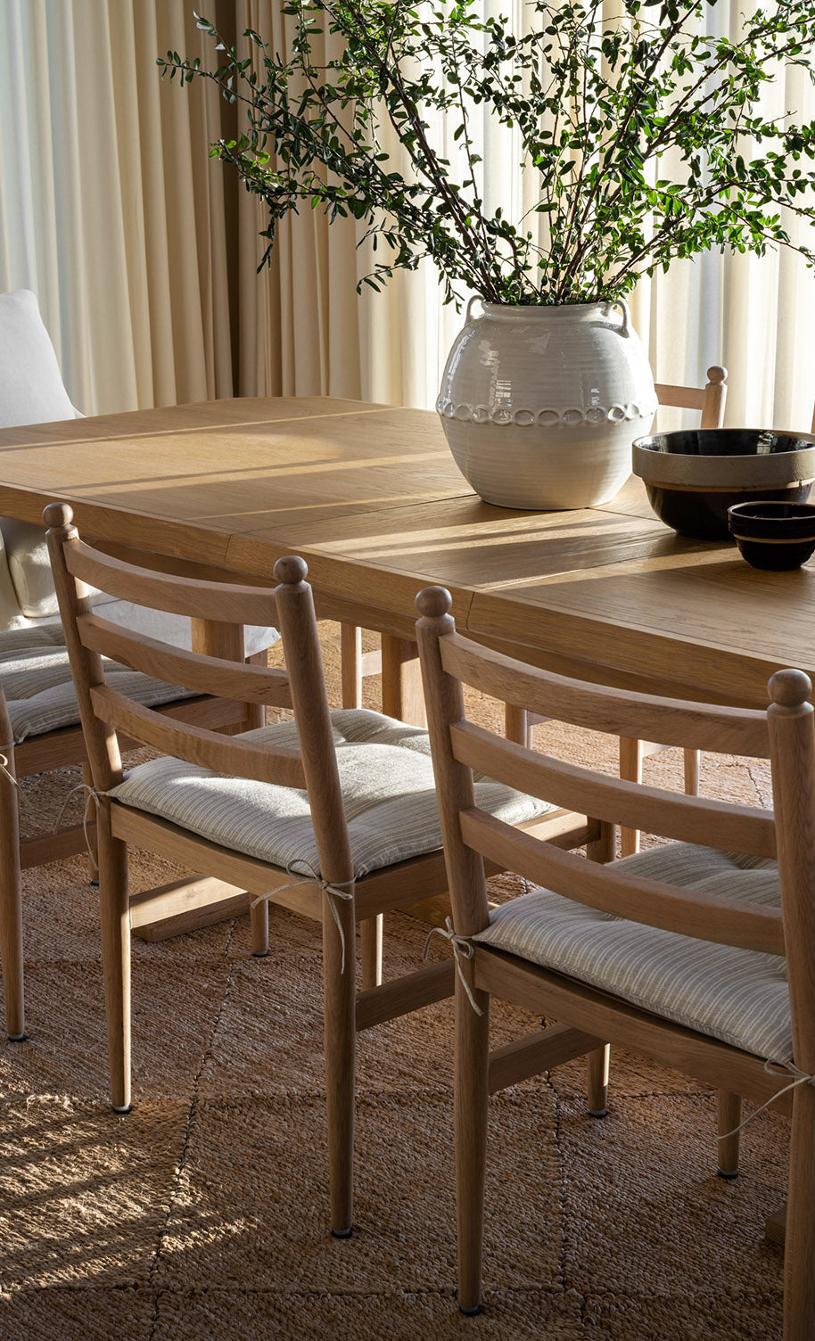 A sunlit wooden dining table with matching chairs, a white ceramic vase holding green branches, and black bowls on a woven rug, with light curtains in the background, by McGee & Co.
