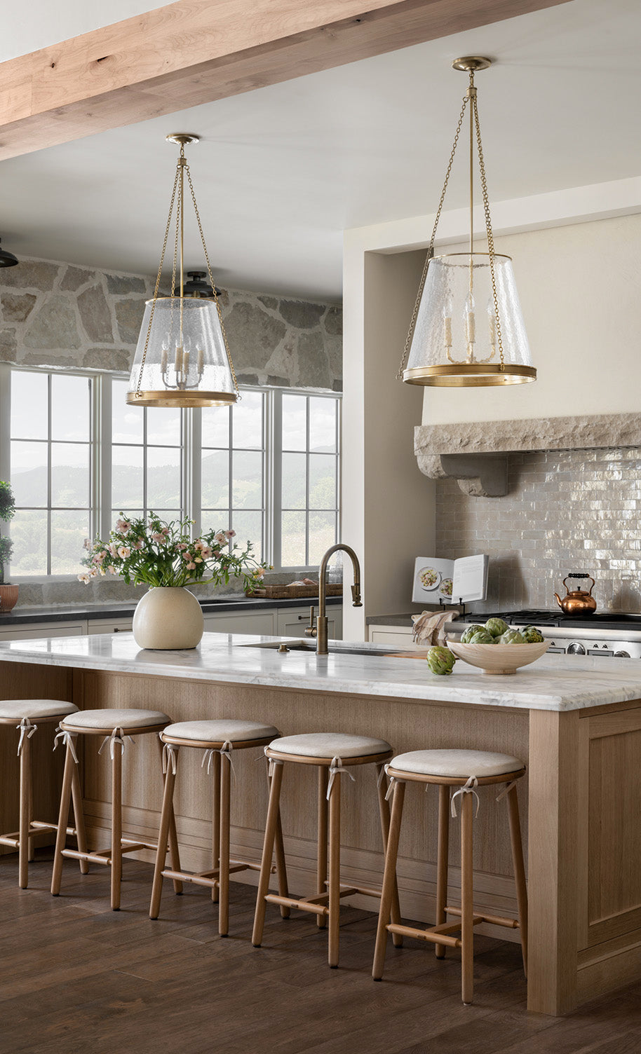A bright, modern kitchen with light wood cabinets, a marble island with four wooden stools, pendant lights, a stone backsplash, large windows, and a vase of flowers on the counter by McGee & Co.