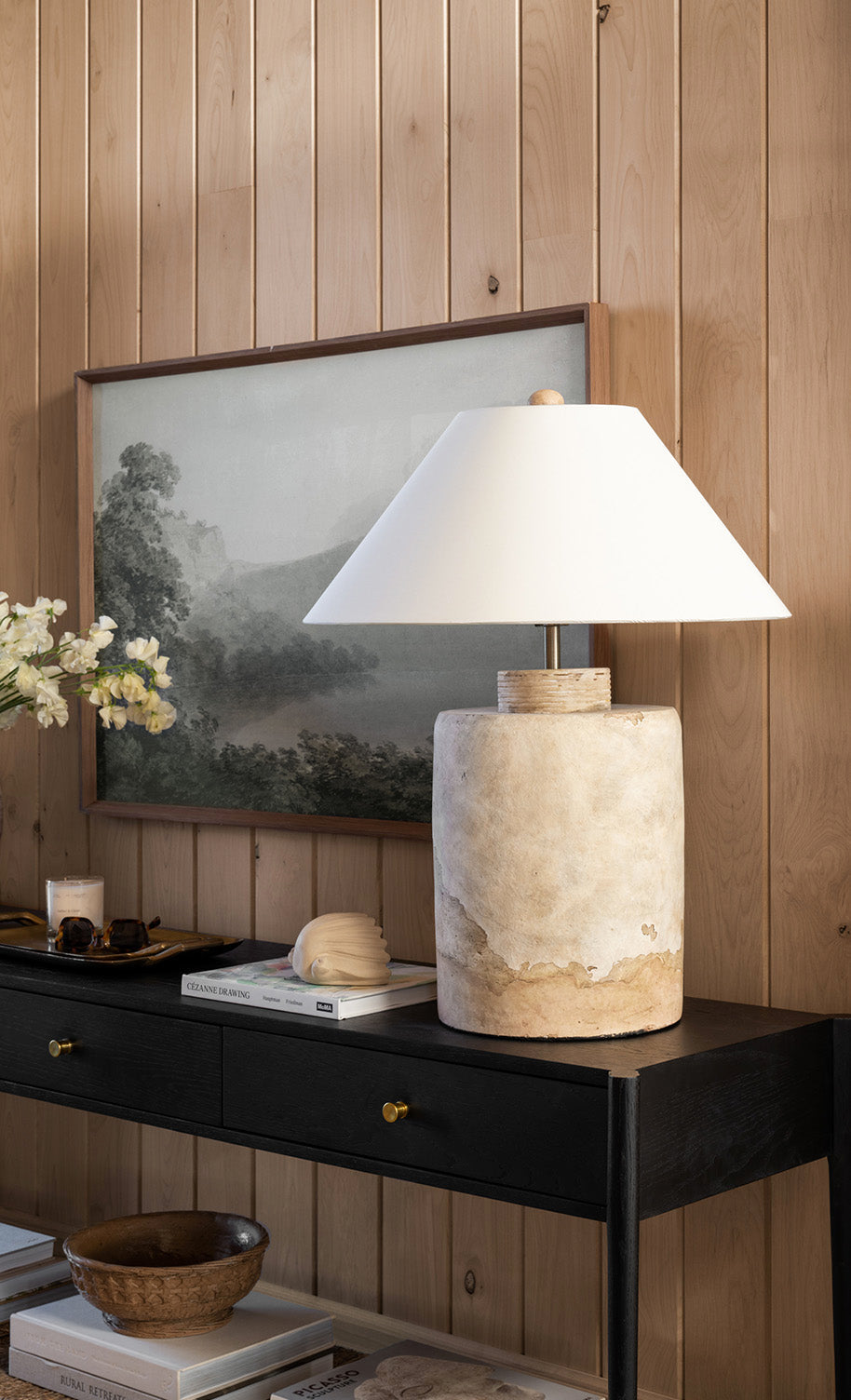 A rustic ceramic lamp with a white shade sits on a black console table, beside stacked books and a white flower arrangement, against a wood-paneled wall with a framed landscape painting, by McGee & Co.