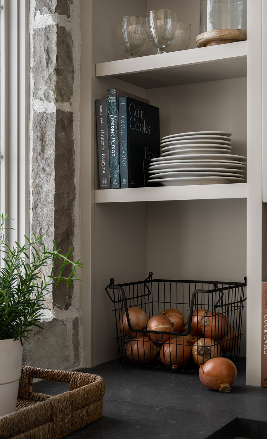 Open shelving in a kitchen with stacked white plates, cookbooks, glassware, a wire basket of onions, a potted plant, and a stone wall beside a window by McGee & Co.