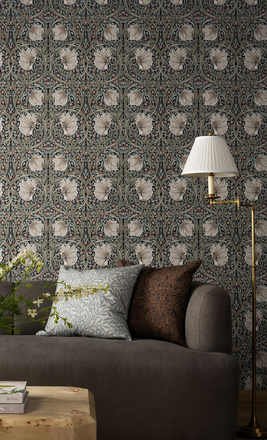A gray sofa with patterned cushions sits in front of a detailed floral wallpaper, next to a wooden table and a gold floor lamp with a white pleated lampshade, by McGee & Co.