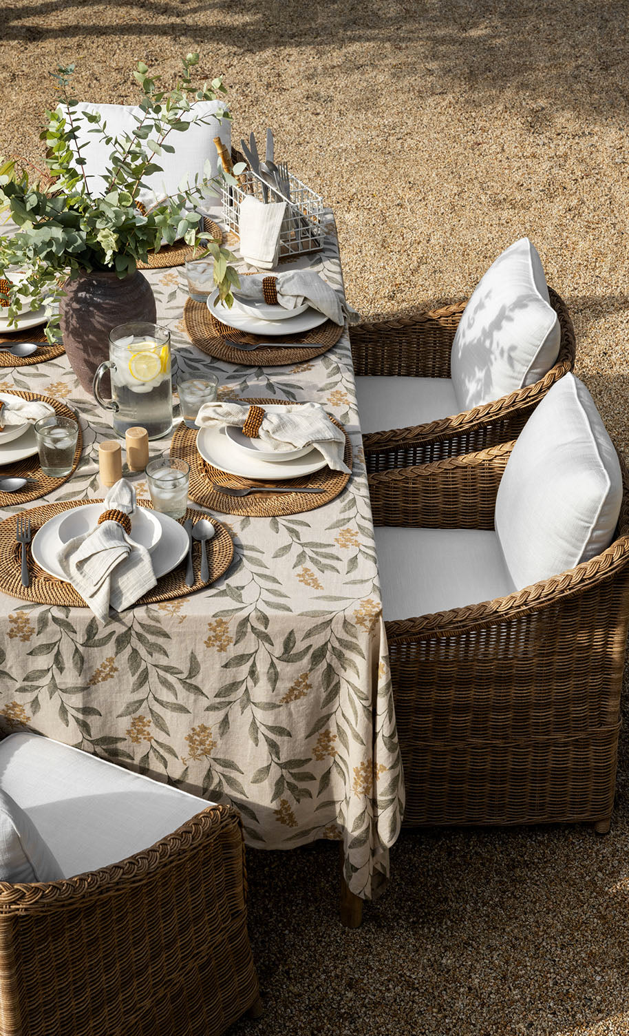 A wicker outdoor dining set with white cushions is arranged around a table covered in a floral tablecloth, set with plates, napkins, glasses, and a jug of lemon water. A vase with greenery is the centerpiece.