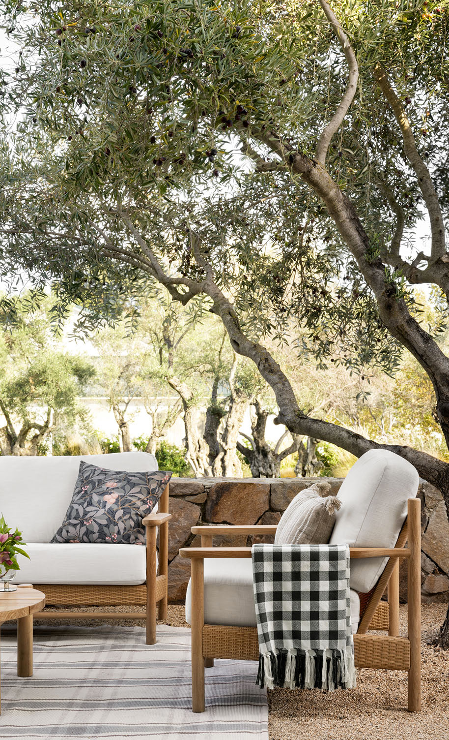 Outdoor seating area with cushioned wooden chairs, decorative pillows, a plaid throw blanket, and a small table, set under leafy trees with a stone wall and greenery in the background.