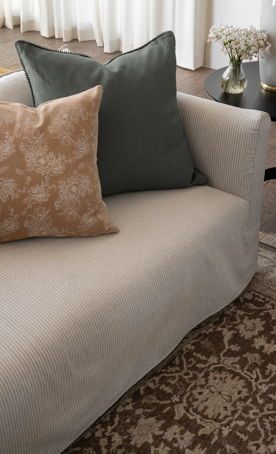 A light gray pinstriped sofa with a green cushion and a beige floral-patterned cushion, next to a round black side table with a vase of flowers, on a patterned rug by McGee & Co.