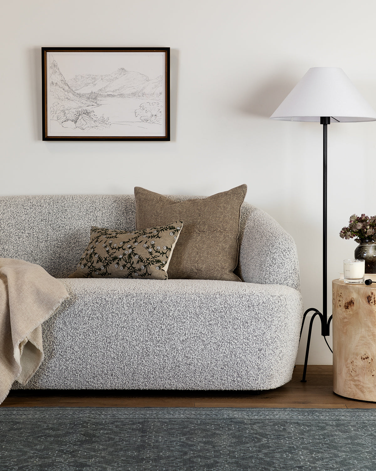 A cozy living room featuring the Makerspalm Byron Settee 68 in textured light gray, two neutral throw pillows, a beige throw blanket, a wood side table with decor accents, a floor lamp, a framed landscape sketch, and a blue-patterned rug—styled with pieces from McGee & Co.