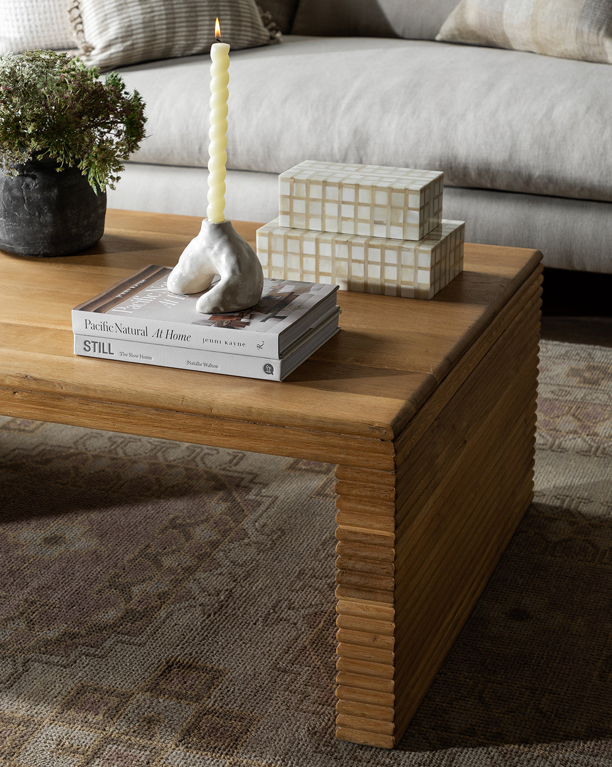 A wooden coffee table with stacked books, a ceramic candle holder, a potted plant, and decorative boxes evokes the Pacific Natural at Home look, atop a patterned rug in front of a light sofa.