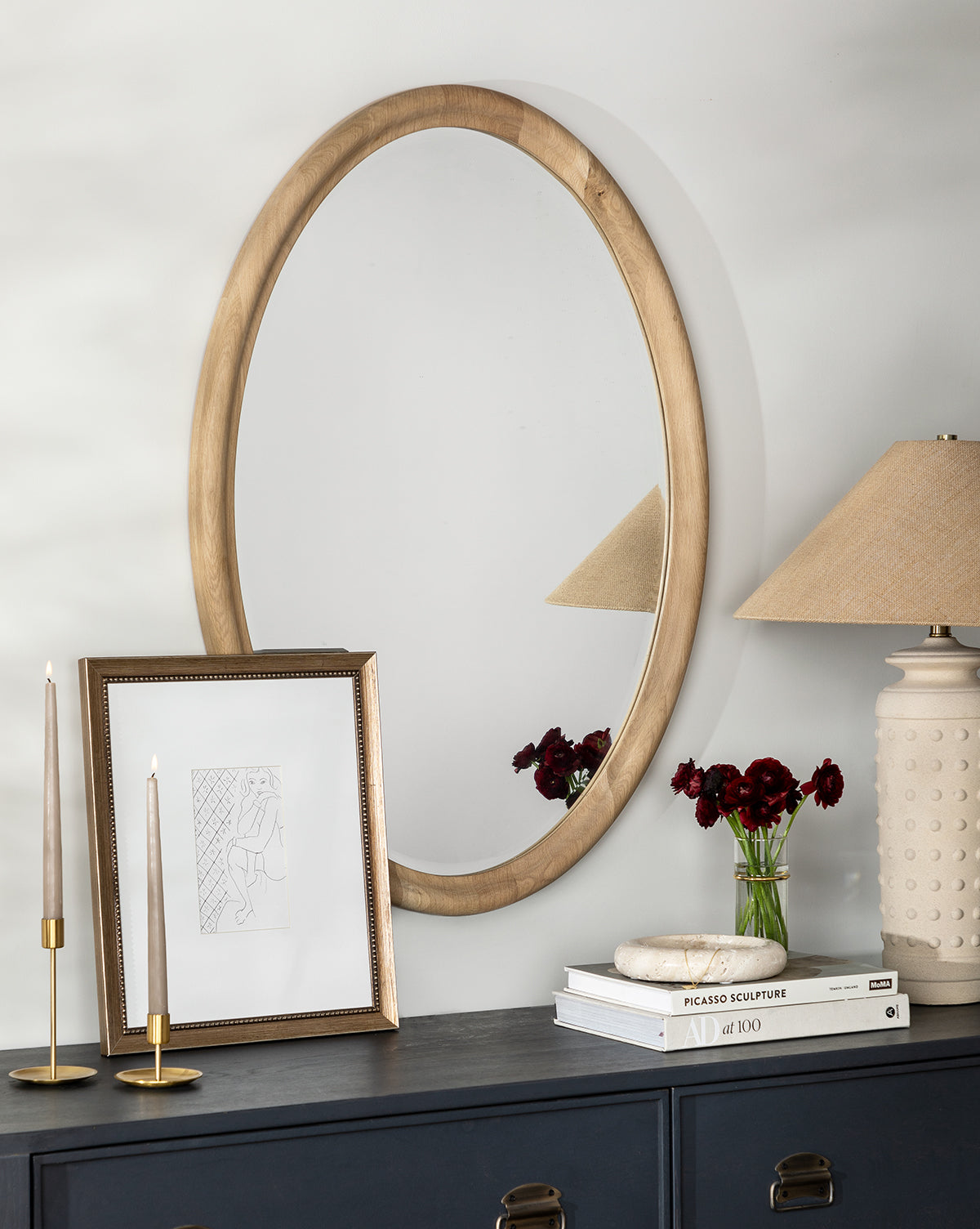 A round mirror with a traditional frame hangs above a dark cabinet topped with a table lamp, the Simple Sketched Woman framed art, candles, a vase of dark red flowers, and stacked books.