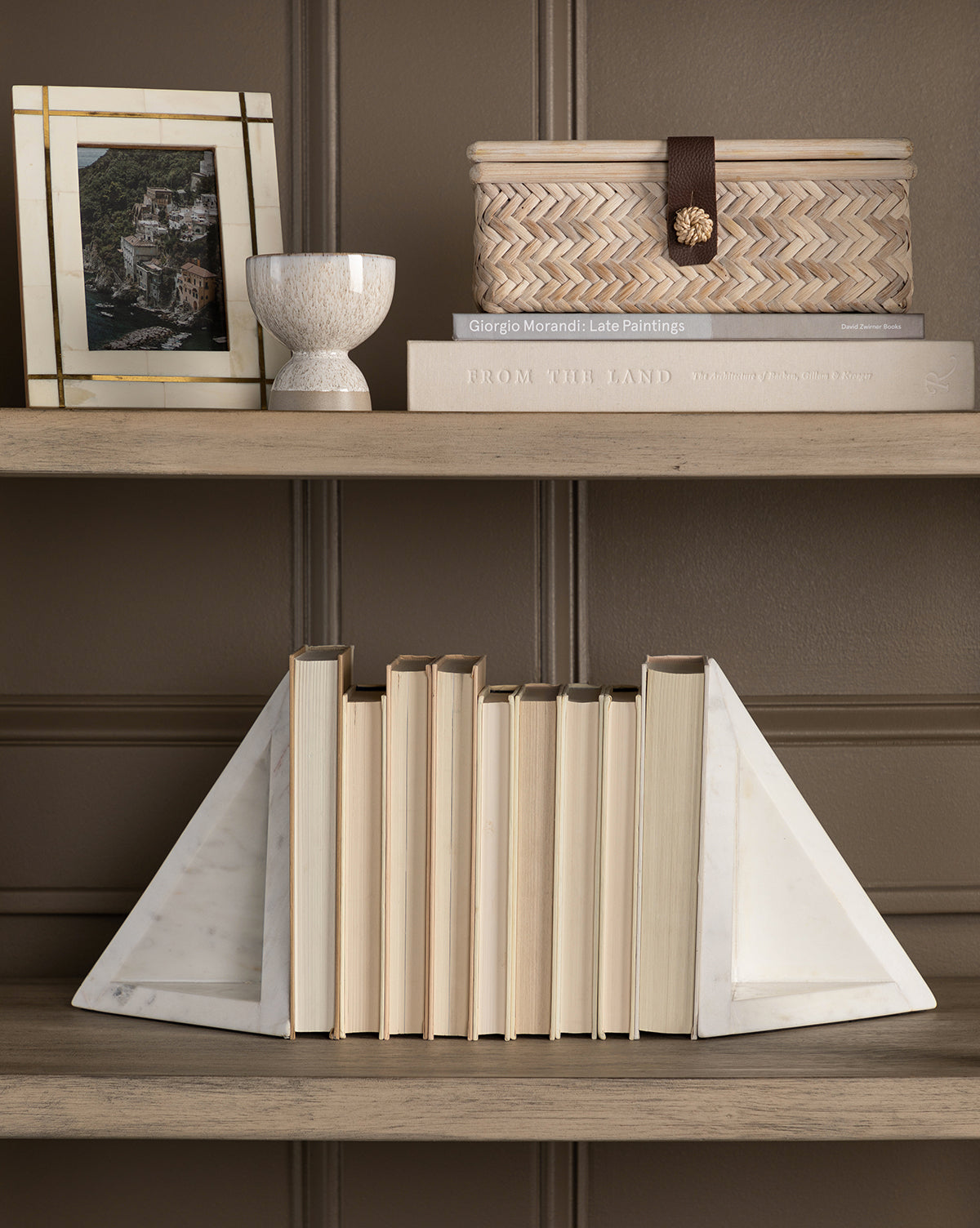 A bookshelf displays upright books with beige spines supported by white marble bookends. Above sits the Winnie Wicker Box, two stacked books, a framed photo, and a small white decorative cup.