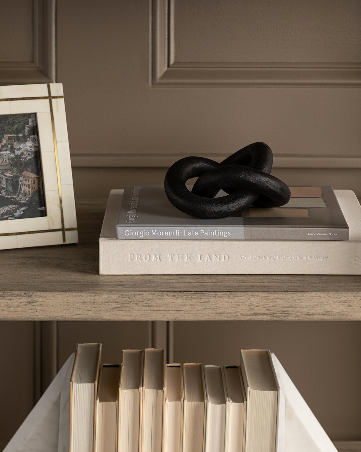 A wooden shelf displays stacked books, including From the Land and Giorgio Morandi: Late Paintings, topped with the Black Infinity Loop metal decor; a framed photo sits beside it with more books below—an elegant modern accent.