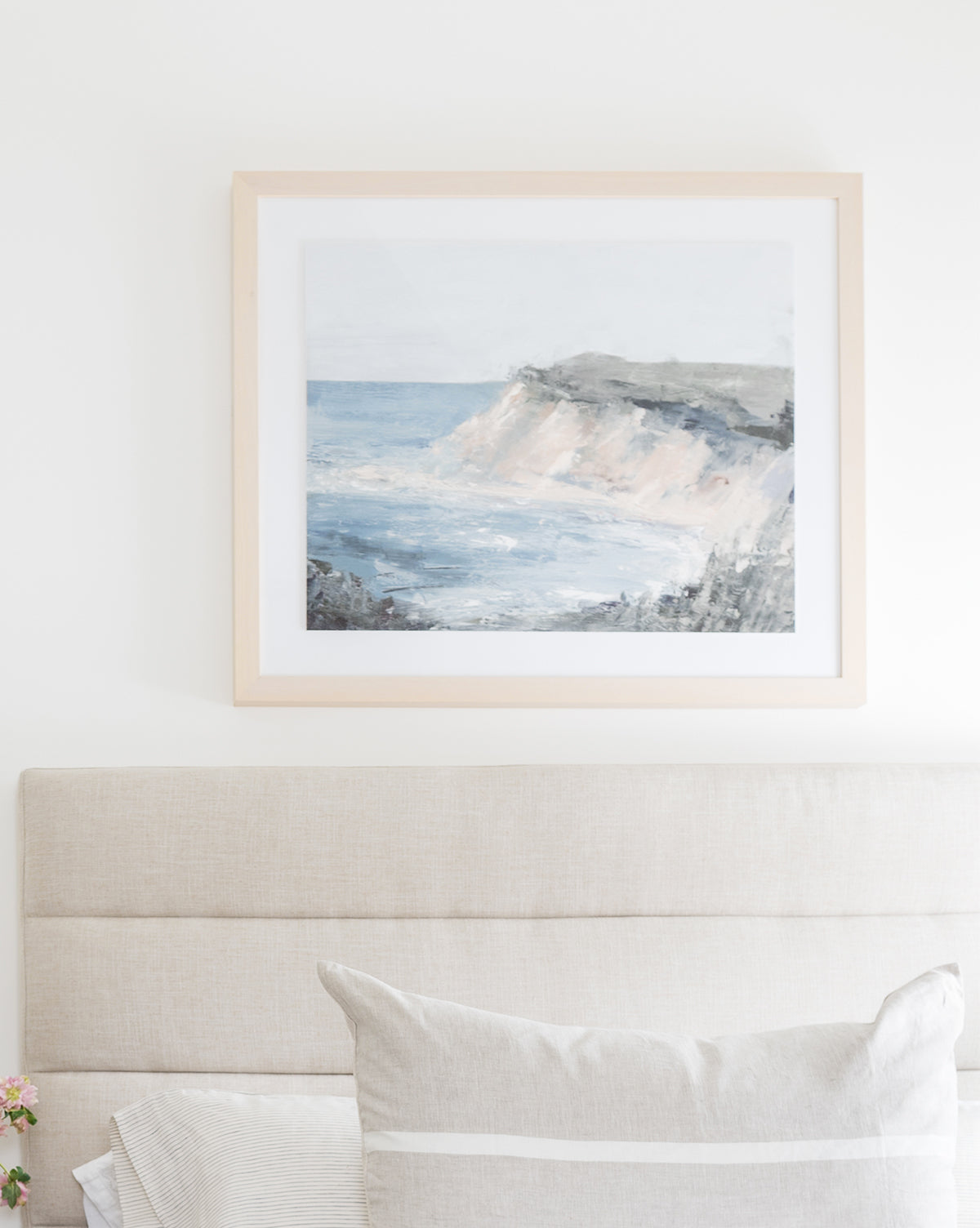 The Shoreside framed painting of a coastal cliff and ocean hangs above a beige upholstered headboard with neatly arranged pillows in this bright, minimalist bedroom.