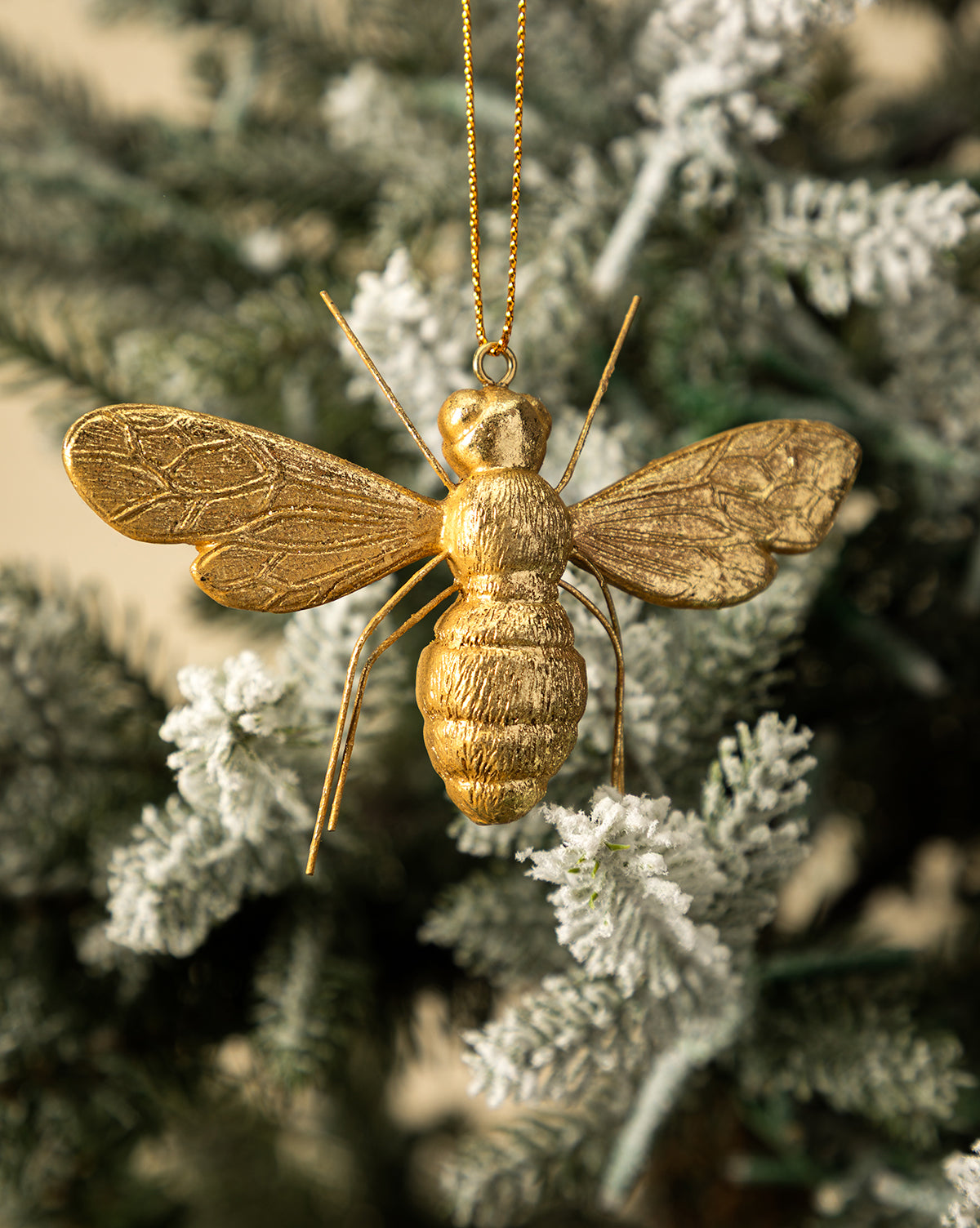 The Golden Bee Ornament, inspired by nature, hangs from a gold string on a snow-frosted holiday tree branch, by McGee & Co.