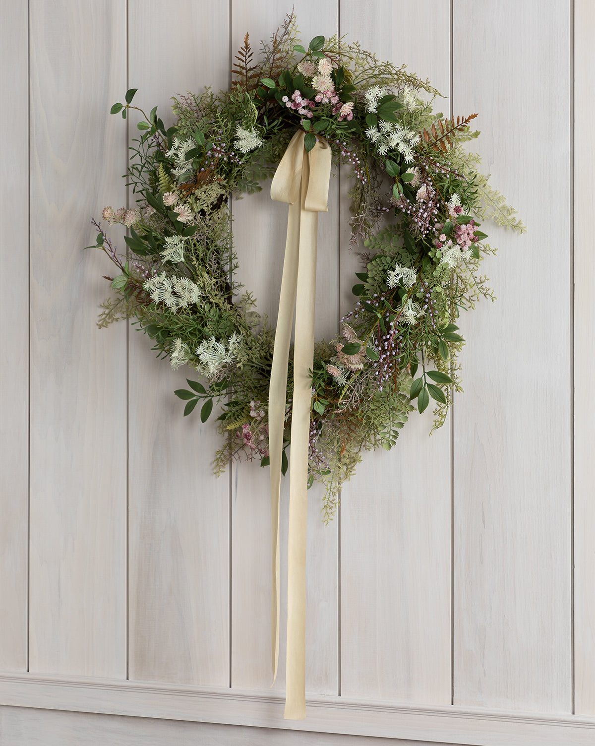 The Faux Herbiflora Wreath, featuring green foliage, pink and white flowers, and ferns, hangs on a light wooden wall. A cream-colored ribbon is tied in a bow at the top, with its ends trailing down this charming faux wreath.