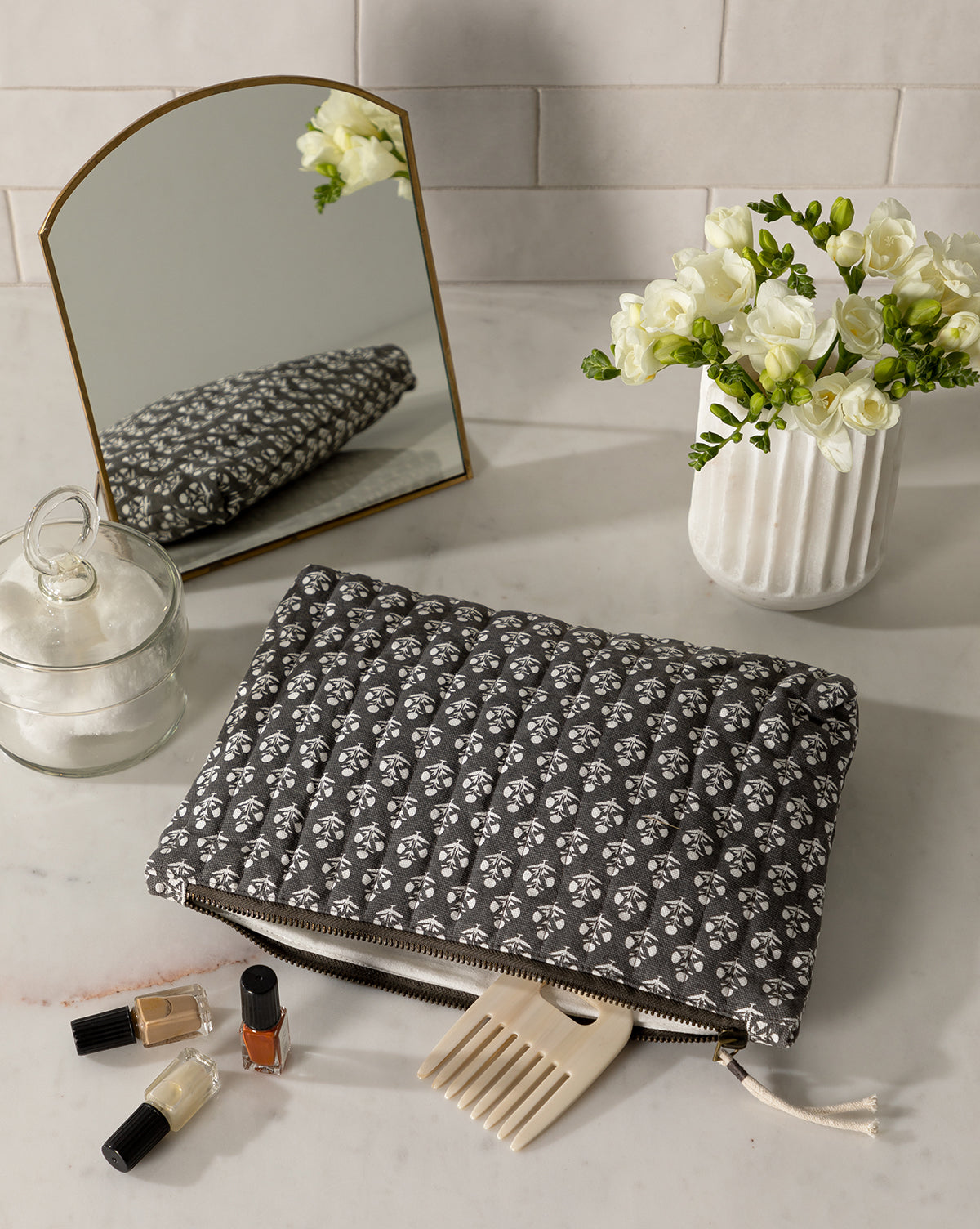 A quilted, floral pouch rests on a marble countertop next to a white vase of flowers, nail polishes, a hair pick, and the Standing Vanity Mirror with a brass-finished frame reflecting the charming display.