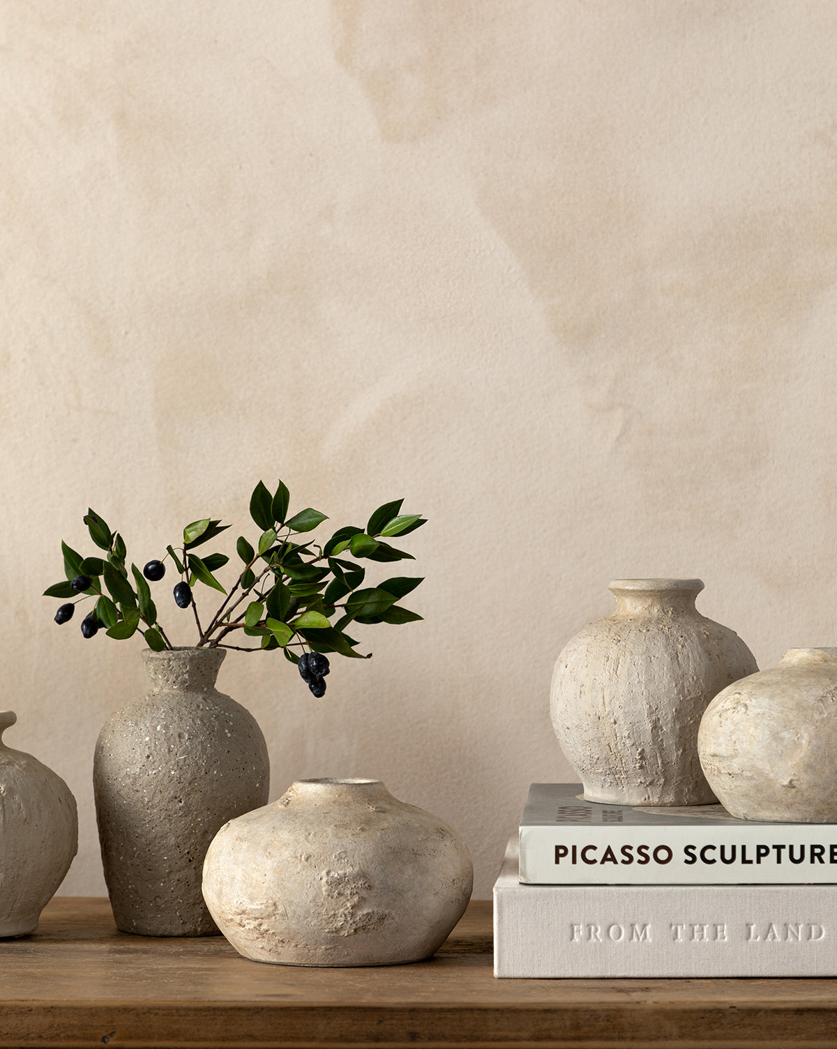 Three textured terracotta Thalia Bud Vases sit on a wooden surface, one filled with green leaves and dark berries, next to two stacked books titled Picasso Sculpture and From The Land. A beige wall forms the background.