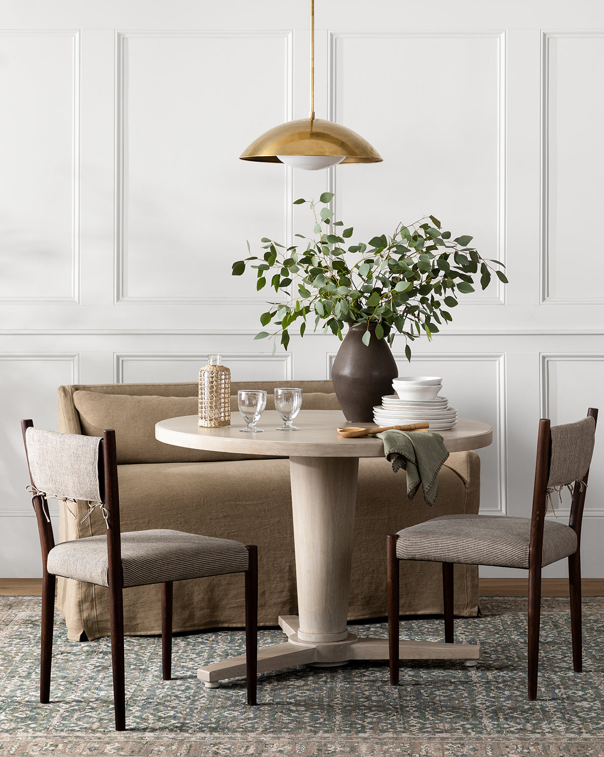 A round light wood dining table with two cushioned chairs and a brown sofa sits on the Marta Hand-Knotted Wool Rug. The table is set with plates, glasses, a vase of greenery, and lit by a gold pendant lamp against a white paneled wall, by McGee & Co.