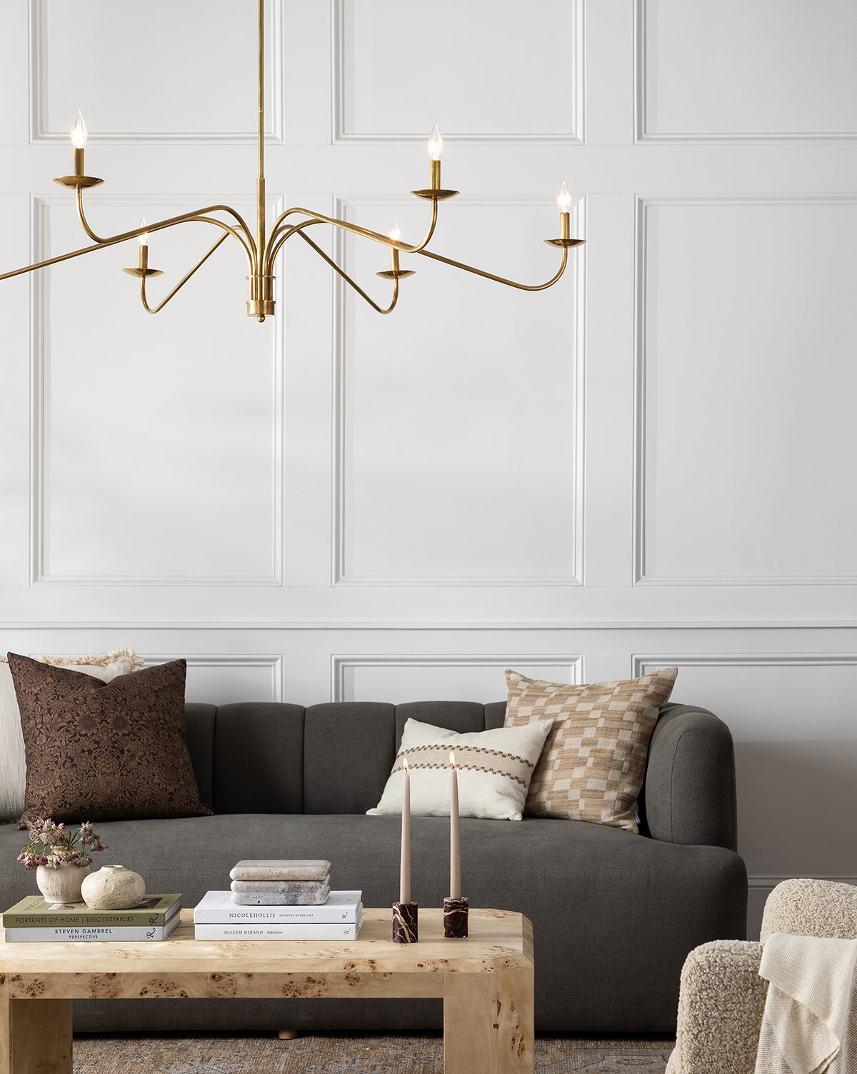 A modern living room featuring the Ines Sofa 90 in gray velvet with patterned pillows, a light wood coffee table with books and candles, and a gold chandelier set against white paneled walls, by McGee & Co.