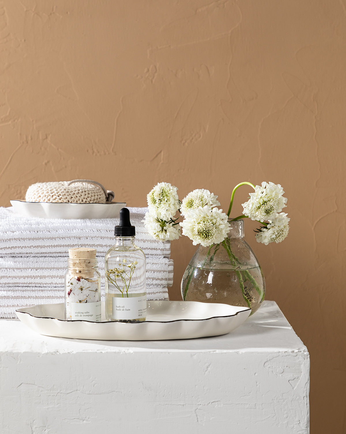 A tray with bath products sits beside a vase of white flowers, while the Taupe Striped Bath Collection—plush, absorbent towels and a washcloth—rests against a beige textured wall.