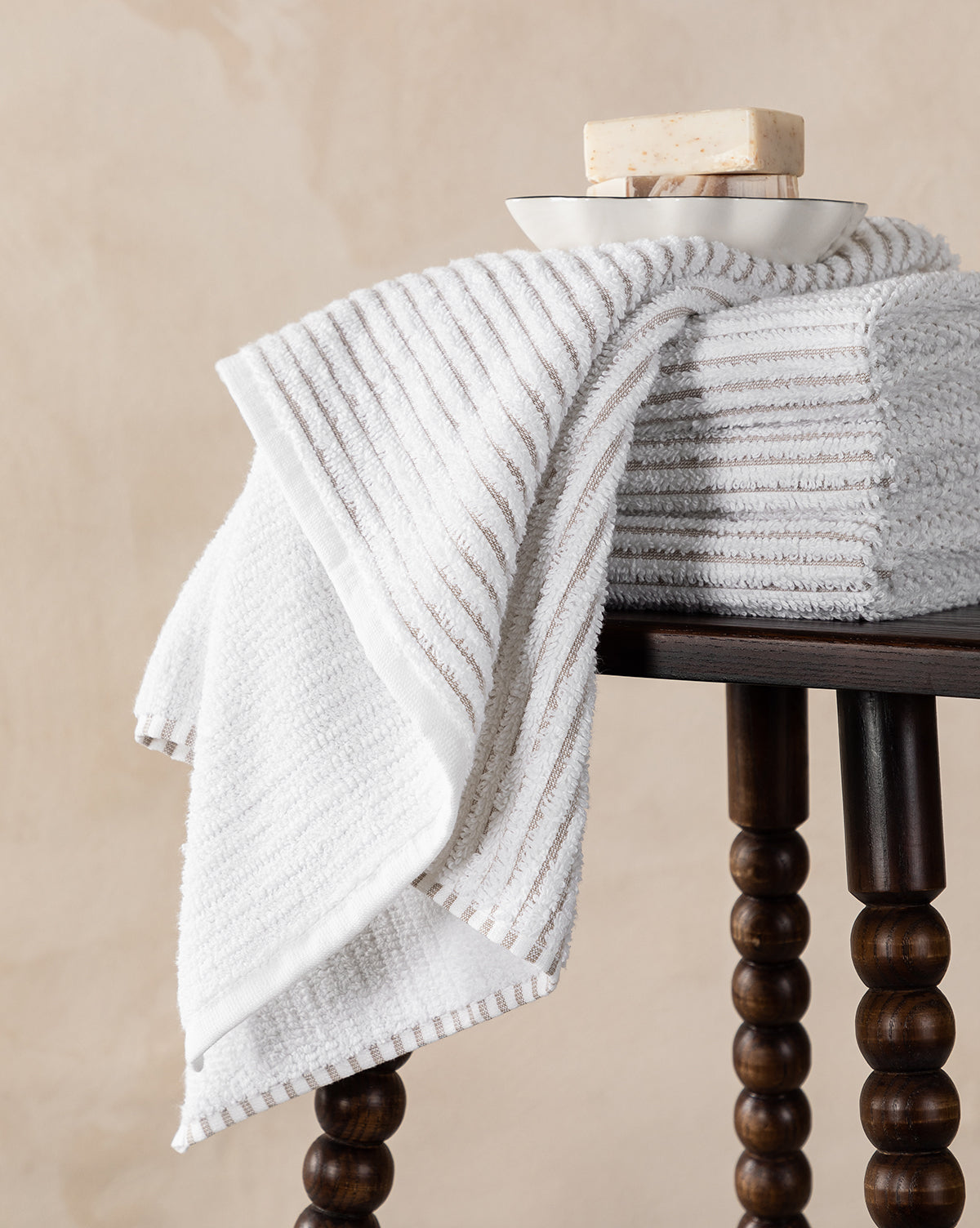A neatly folded stack from the Taupe Striped Bath Collection sits on a dark wooden stool with a bar of soap in a small dish on top; one absorbent towel hangs over the edge.