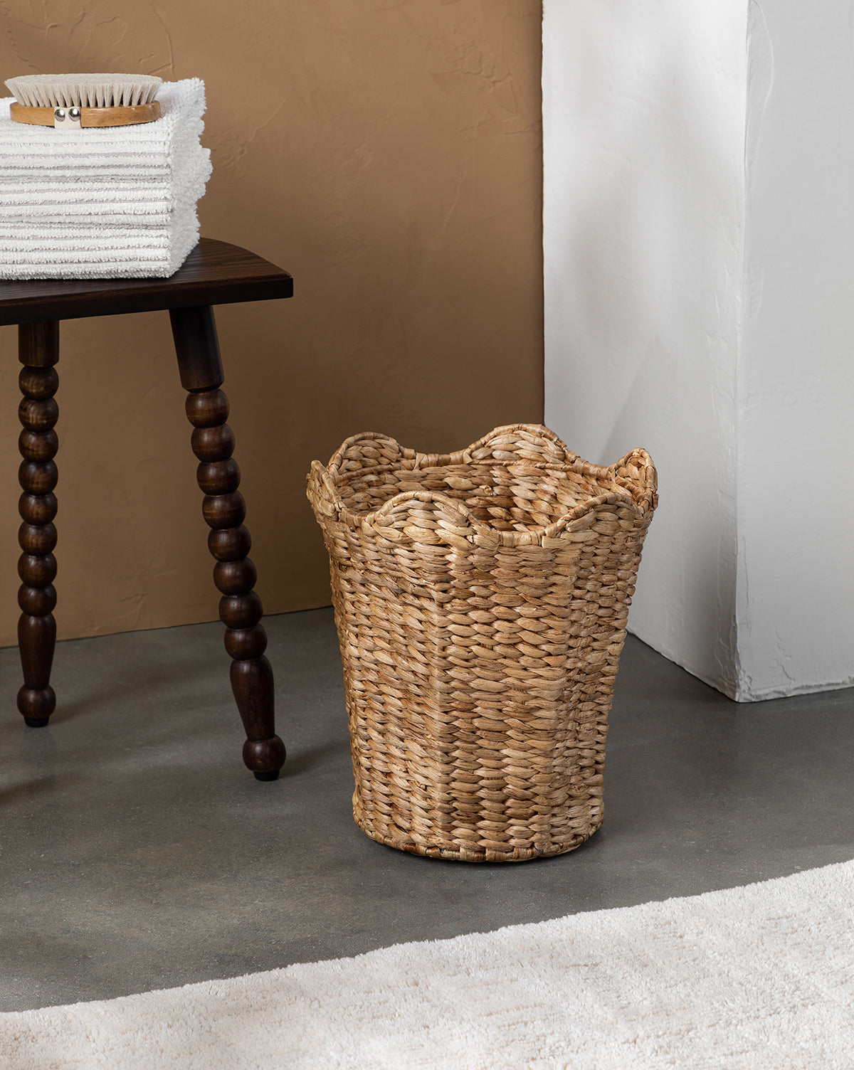 The Gray Striped Bath Collection is displayed by a woven wicker basket on a gray floor, next to a wooden table topped with stacked towels and a brush, against tan and white walls, with part of a white rug in view.
