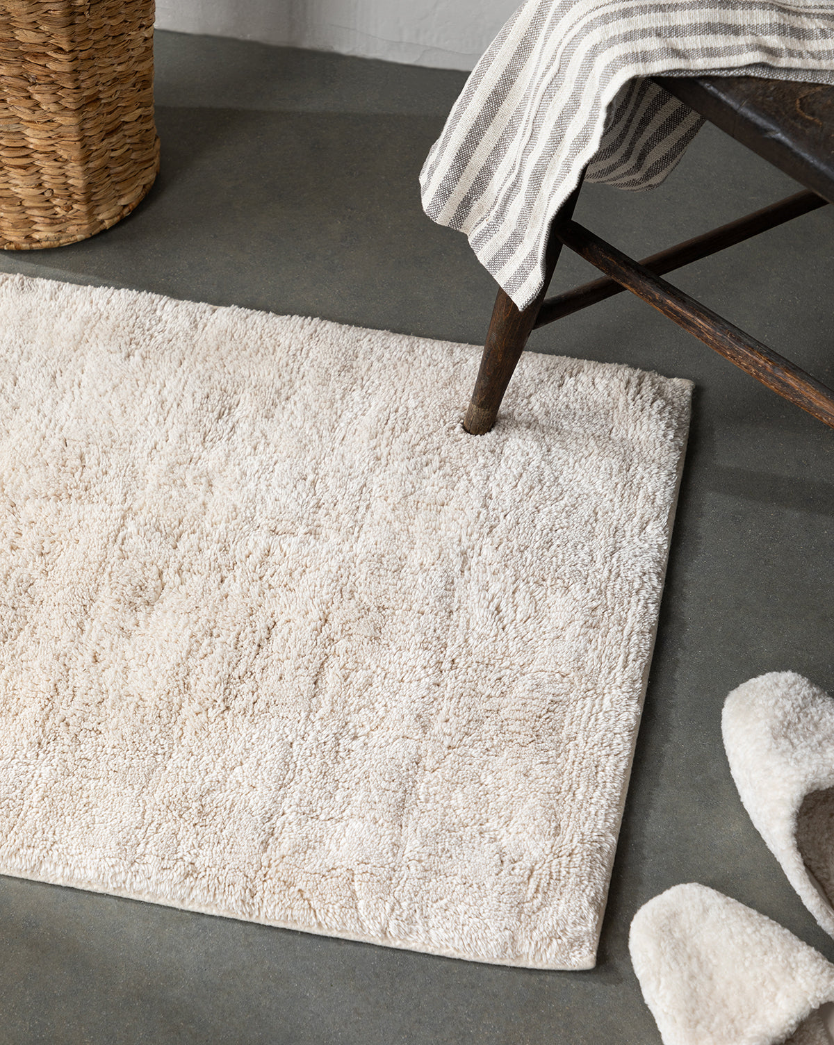 The Callen Bath Mat in cream sits on a gray floor next to fluffy slippers, a striped towel draped over a wooden bench, and a woven basket.