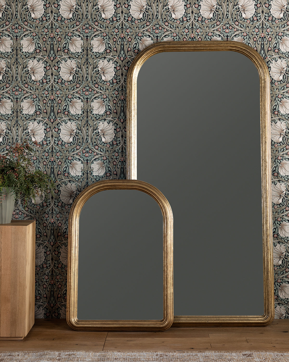 Two Giada Floor Mirrors with elegant frames lean against a wall with floral wallpaper. Nearby, a wooden cabinet topped with a glass vase of greenery sits on a rug-covered floor, by McGee & Co.
