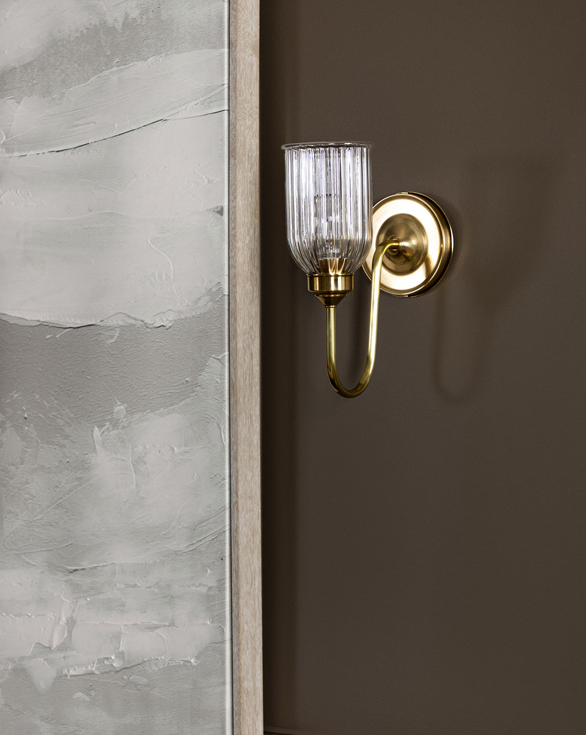 A Makerspalm Clyde Sconce in burnished bronze with a reeded glass shade is mounted on a brown wall beside a textured, light gray painted surface. McGee & Co.