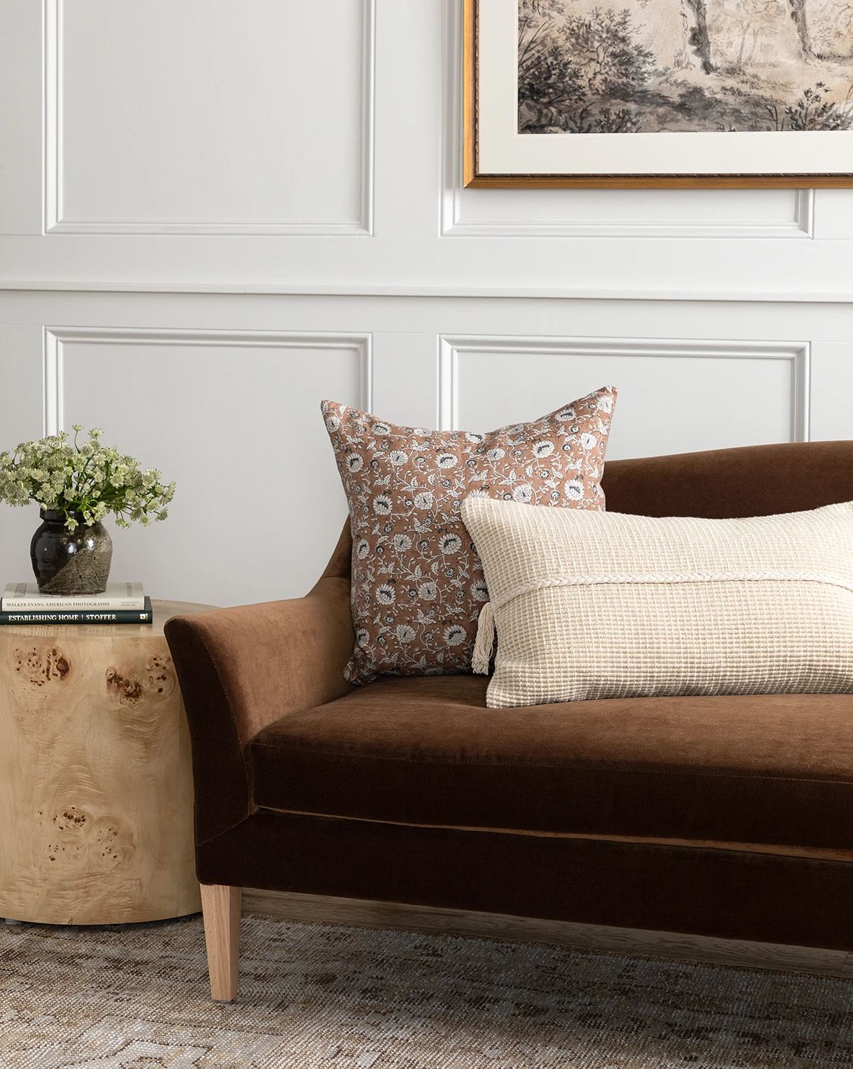 The Prudence Sofa 78 in brown velvet features premium fabric and two accent pillows, paired with a round wooden side table holding a vase of flowers and books, set against a white paneled wall with framed artwork above, by McGee & Co.