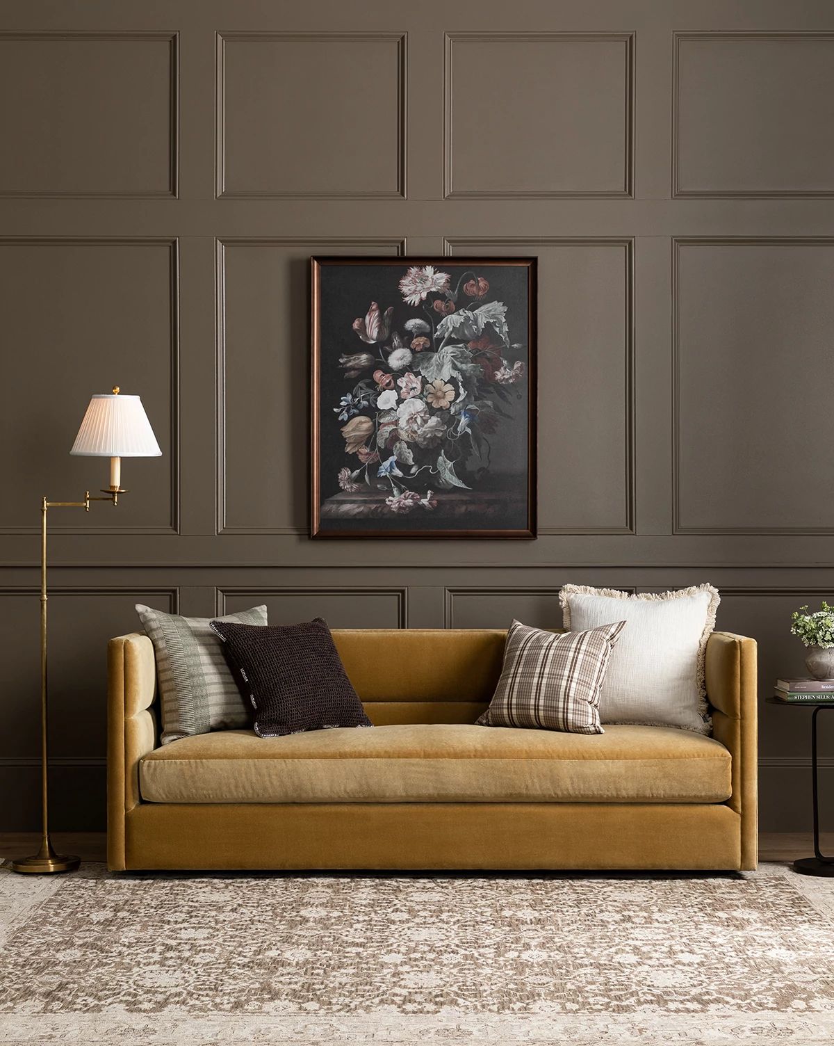 A Claremont Sofa 84 in mustard yellow, accented with assorted pillows, sits against a brown paneled wall under a floral painting. A brass floor lamp and small side table complete this cozy, elegant living room setting, by McGee & Co.
