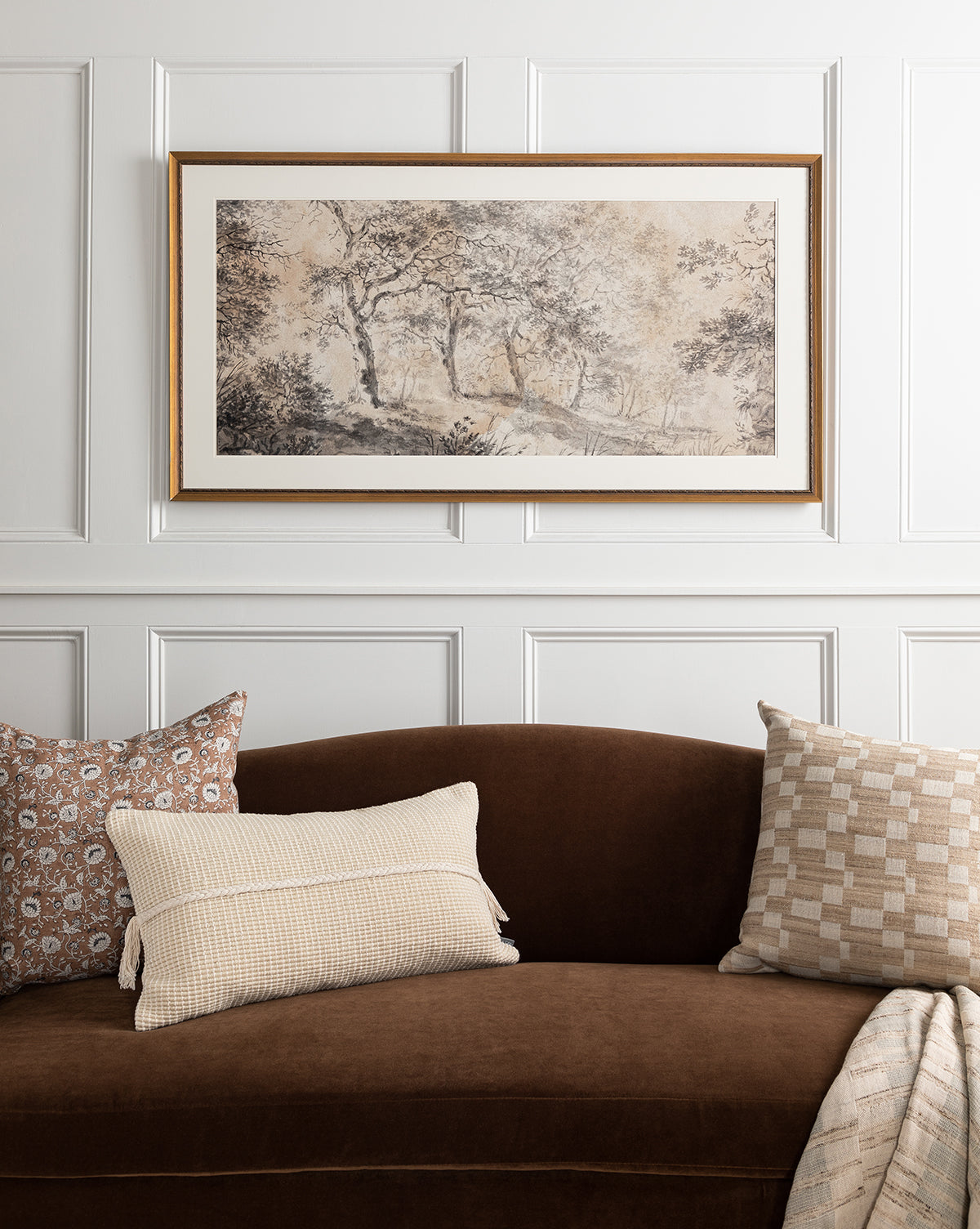 A brown velvet sofa with three neutral patterned pillows sits against a white paneled wall, topped with the Dutch Landscape Study giclee print in an antique gold frame, showcasing muted-toned trees and nature.