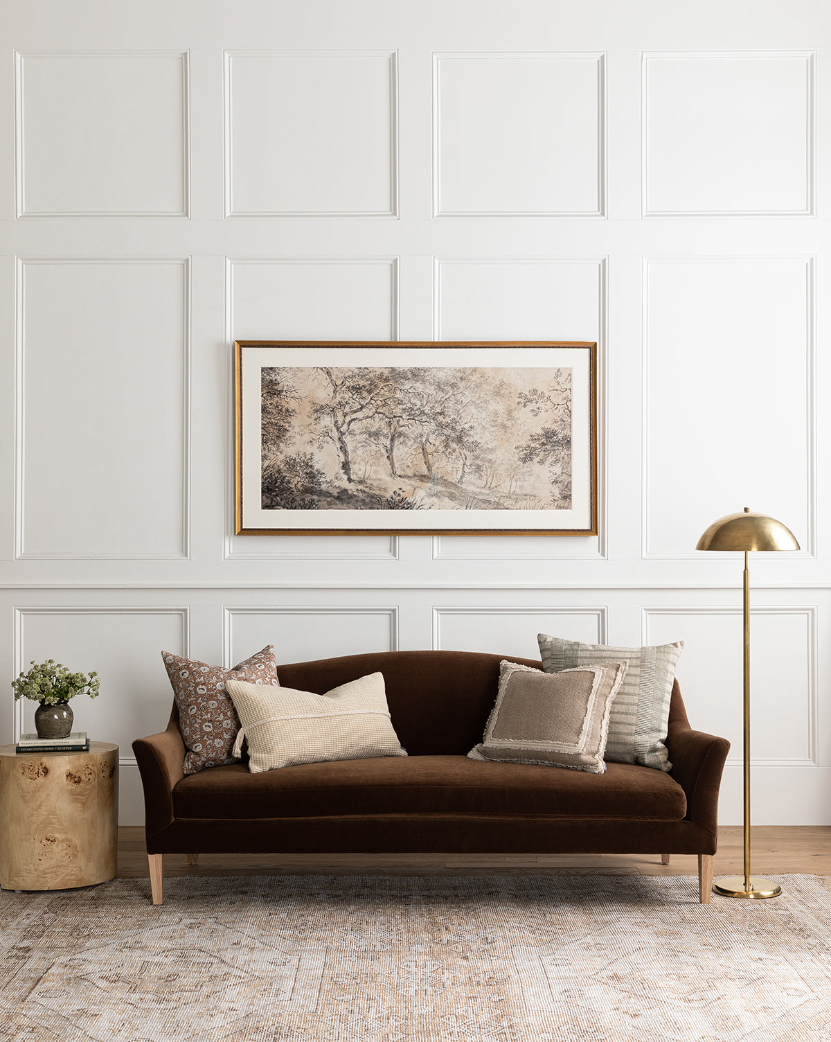 A modern living room featuring a dark brown velvet sofa with neutral throw pillows, a round wooden side table with a plant, the Aldorno Floor Lamp for elegant lighting, and framed landscape art on a white paneled wall, by McGee & Co.