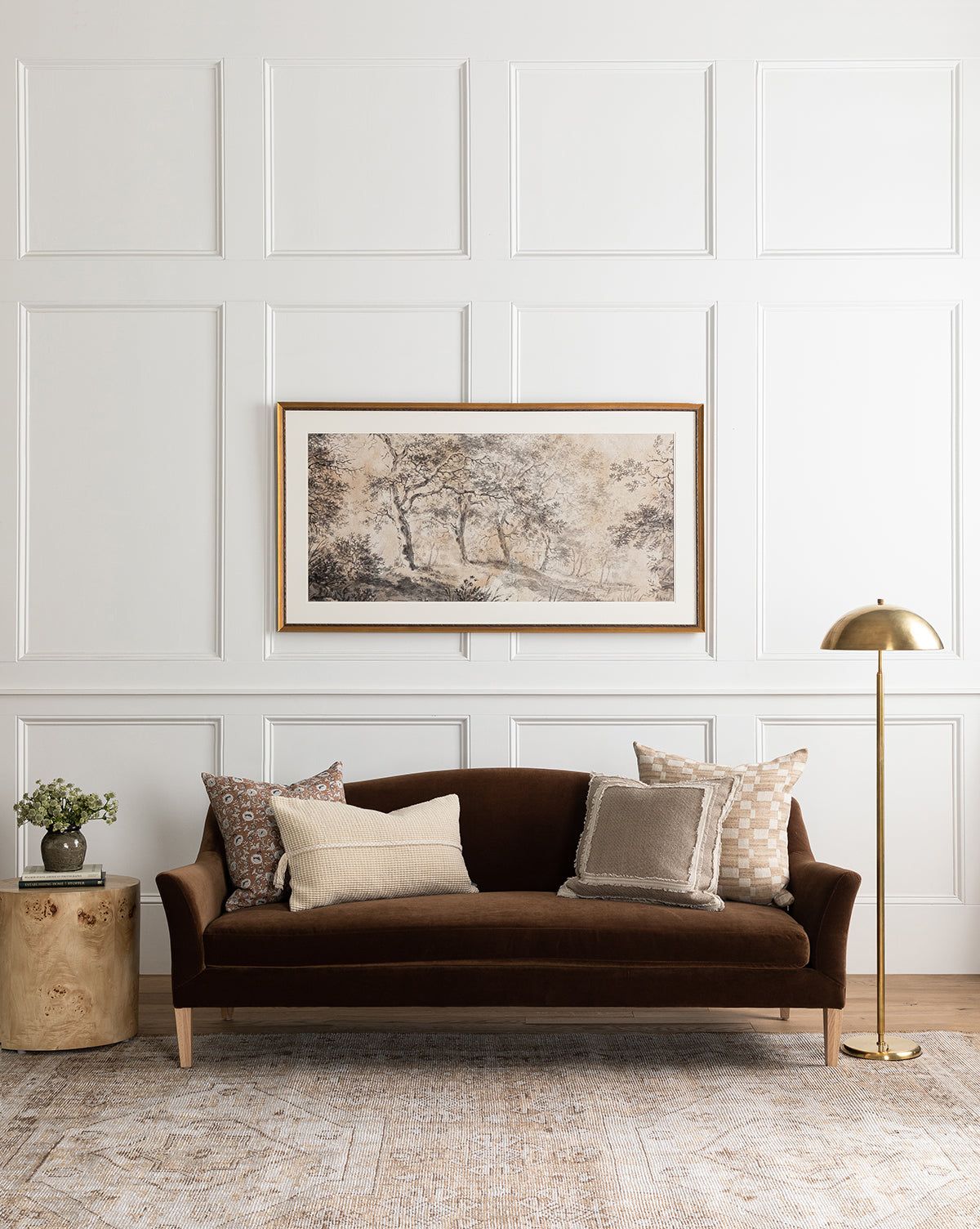 A stylish living room features the Makerspalm Prudence Sofa 78” in dark brown, neutral throw pillows, a round side table with a small plant, a gold floor lamp, and framed landscape art on a white paneled wall—finished with touches from McGee & Co.