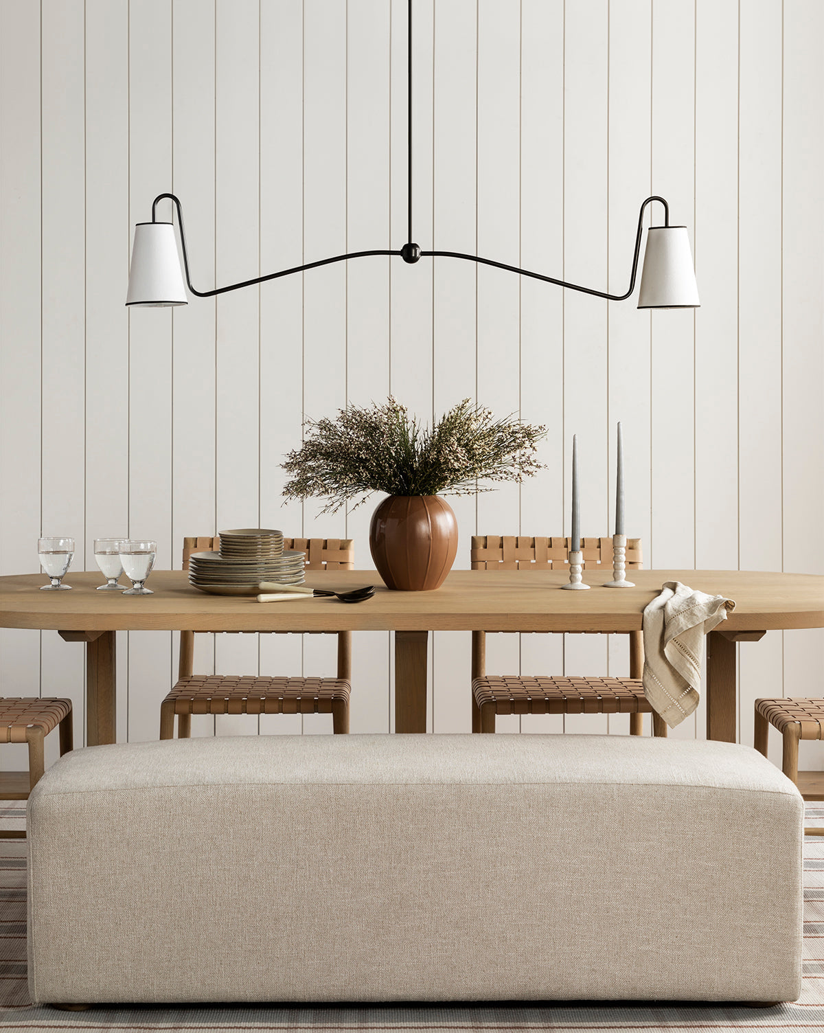 A minimalist dining room showcases a light wood table set with plates, glasses, and candles, a brown vase with greenery, woven chairs, a beige bench, and elegant lighting from the Makerspalm Jonas Linear Chandelier above. McGee & Co.