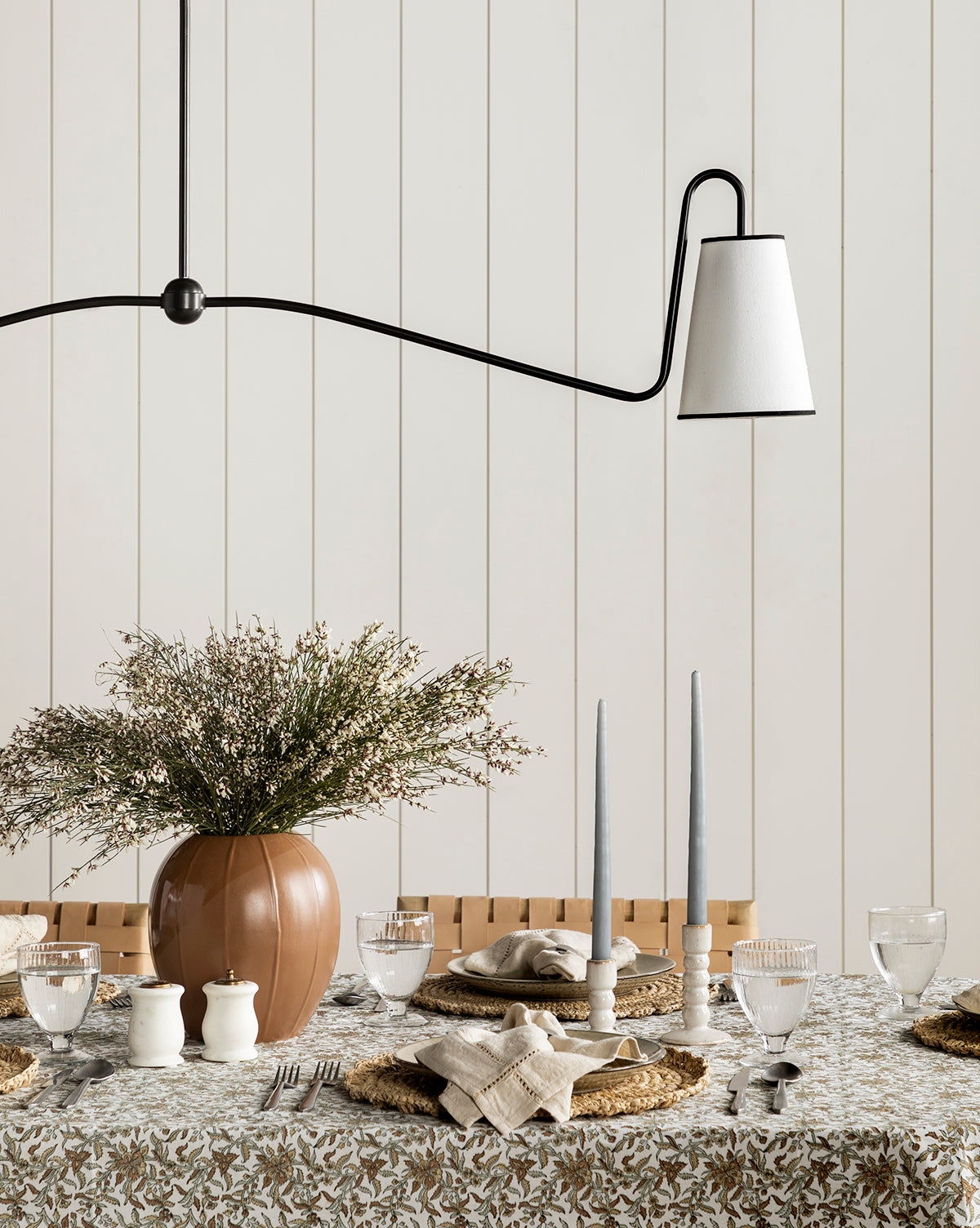 A dining table set with plates, glasses, napkins, a large vase of flowers, and candles in the Mae Ceramic Taper Holder sits beneath a modern black-and-white lamp against a white paneled wall.