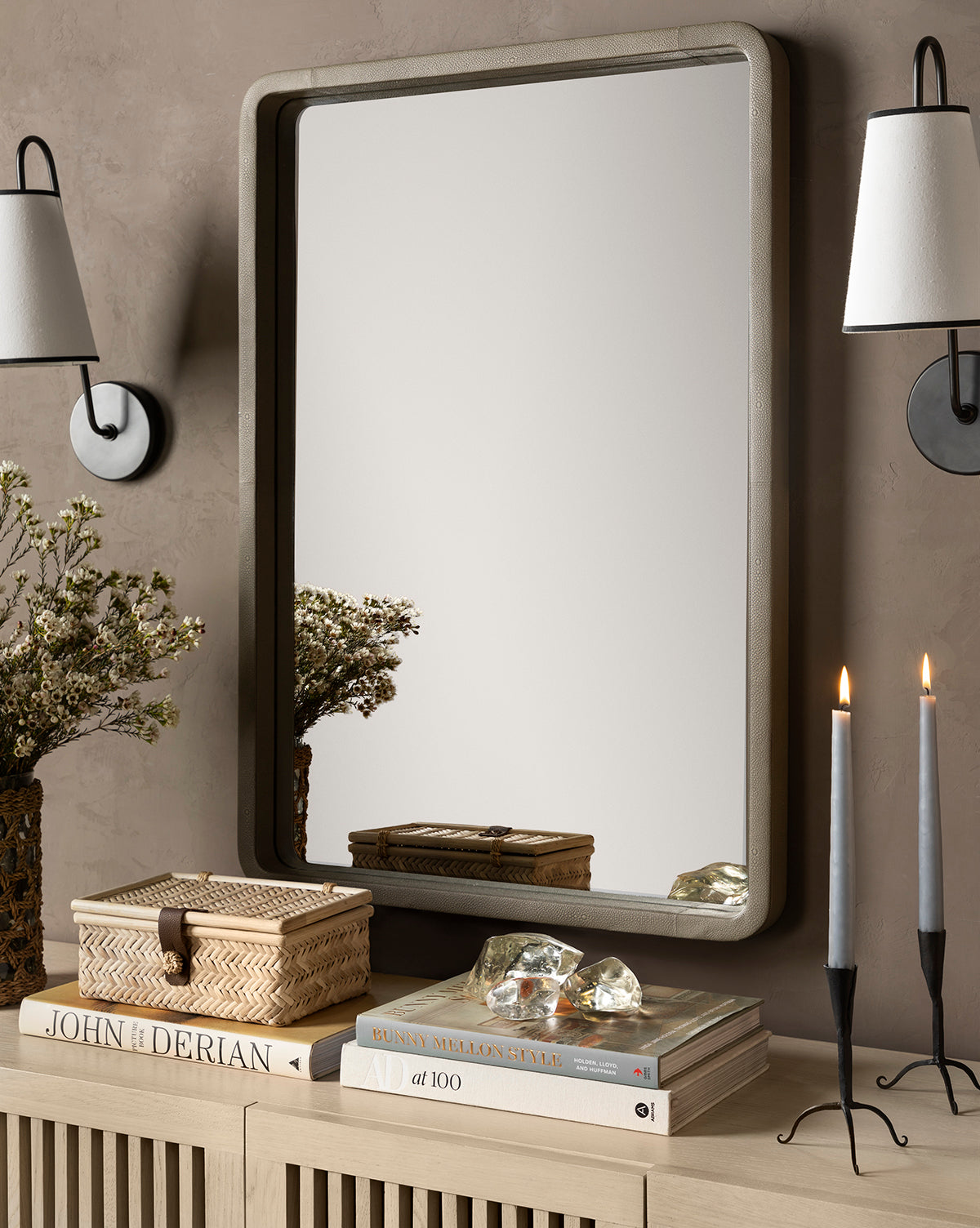 A neutral console table with stacked books, the Winnie Wicker Box, a crystal, and two lit gray candles sits beneath a wall mirror; vases of flowers and wall sconces flank the mirror.