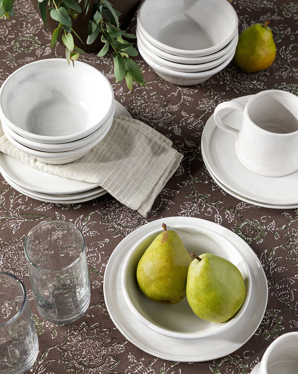 A table set with stacked white bowls, plates, a mug, the Aiden Side Plate, a beige napkin, two pears on a white plate, clear glasses, and green leaves on a brown patterned tablecloth by McGee & Co.