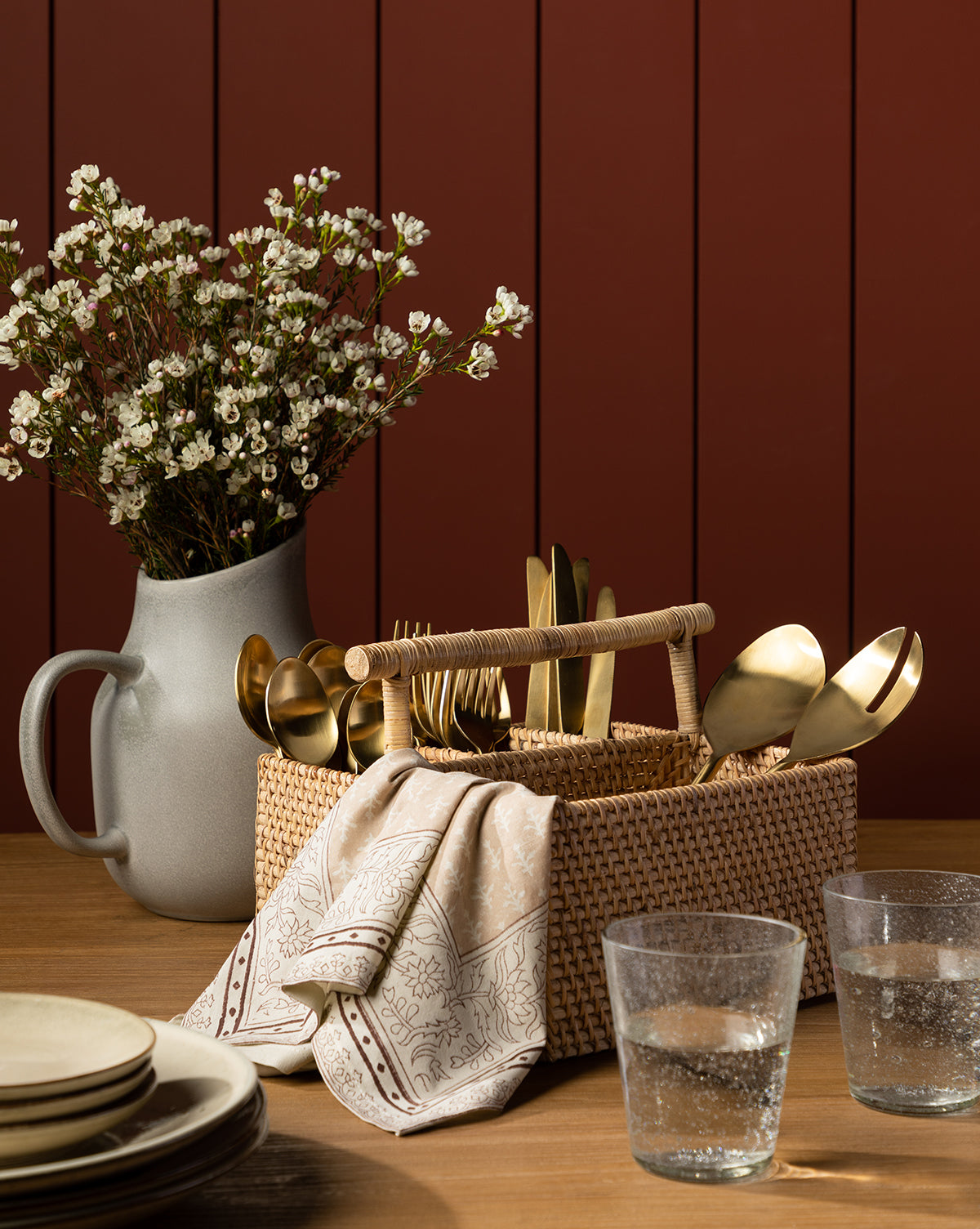 A woven basket with gold-toned flatware and a napkin sits on a wooden table beside two glasses of water, stacked plates, and the Levi Stoneware Pitcher. In the background, a gray vase with white flowers stands against a red-paneled wall, by McGee & Co.