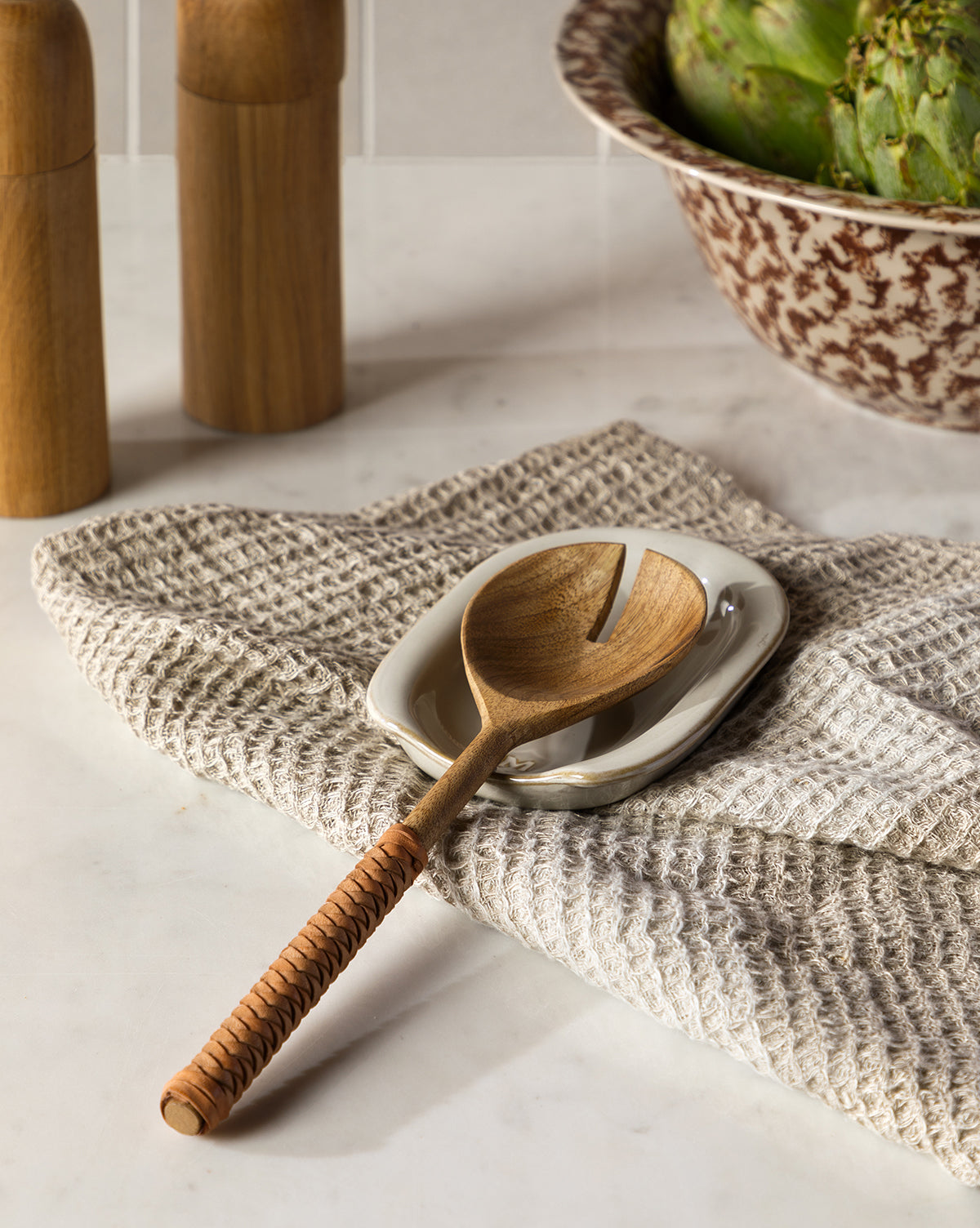 A wooden serving spoon with a woven handle rests on a small white dish and the Bunton Waffle Tea Towel. Wooden salt and pepper mills and a bowl of green artichokes sit in the background, by McGee & Co.
