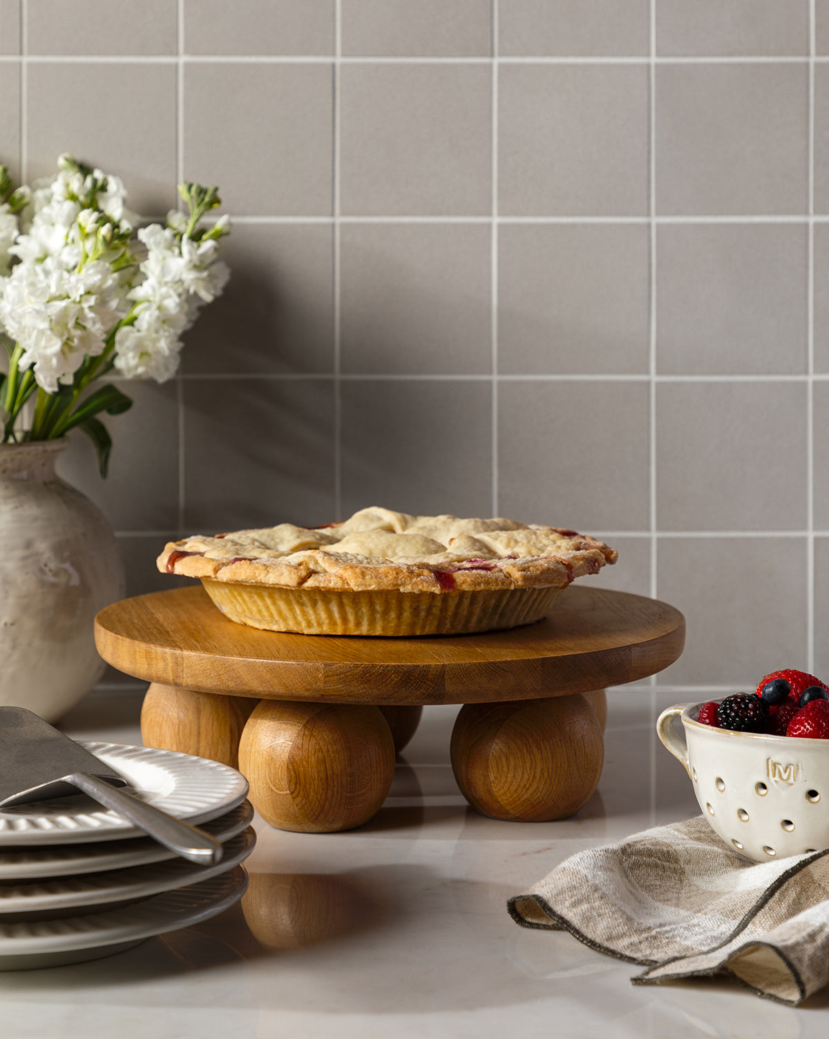 A homemade pie rests on the Bronx Oak Pedestal atop a kitchen counter, surrounded by plates, a pie server, a bowl of berries, a beige napkin, and a vase of white flowers in the background, by McGee & Co.
