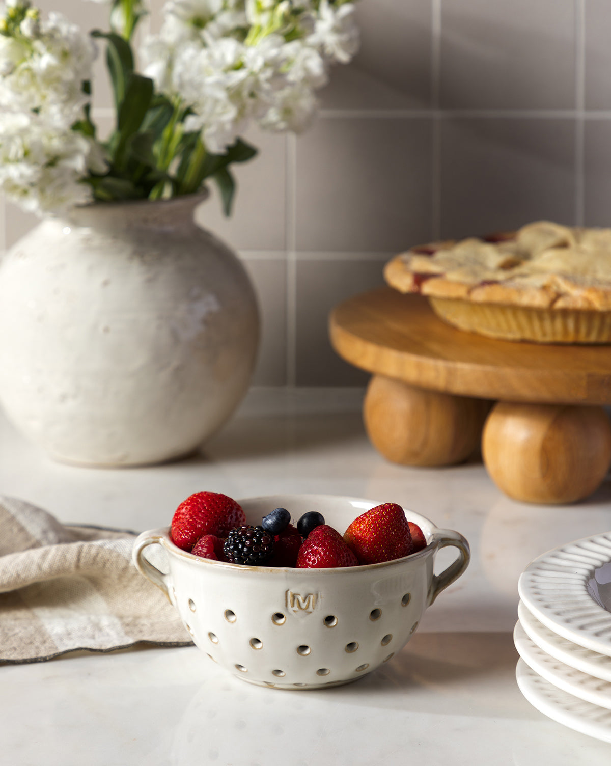 A white Glazed Berry Colander brimming with strawberries, blackberries, and blueberries sits on a kitchen counter next to stacked serveware, with a pie and white flowers enhancing the kitchen decor in the background, by McGee & Co.