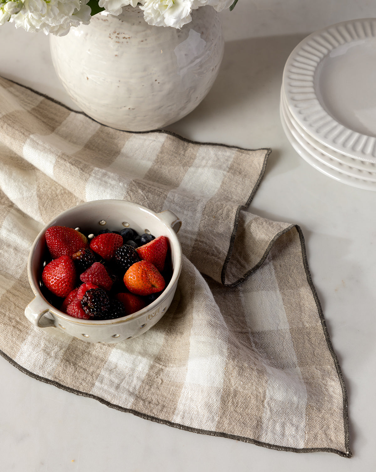 The Charlie Check Tea Towel in beige holds a white ceramic bowl of strawberries, blackberries, and blueberries beside stacked white plates and a white vase of flowers on a marble surface, by McGee & Co.