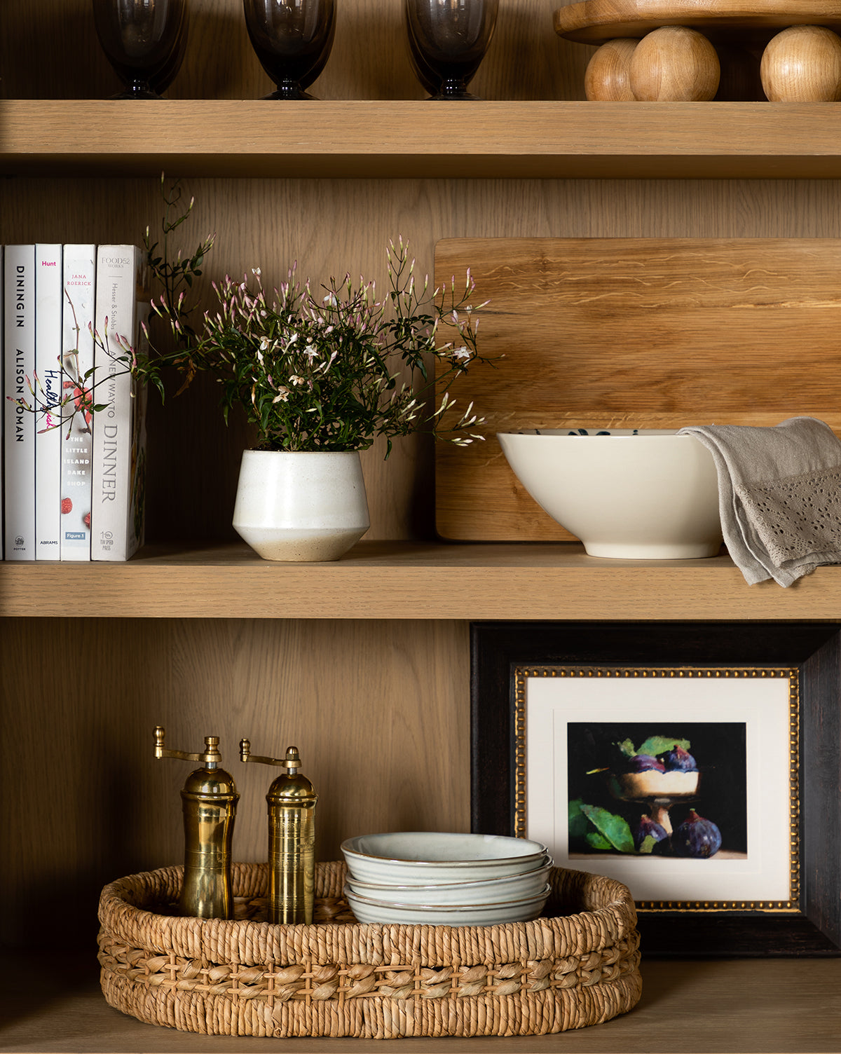 A wooden shelf displays books, potted flowers, a bowl with cloth, the Figs Still Life framed photograph, brass salt and pepper mills, stacked dishes in a woven tray, and black and gold wood decor pieces.