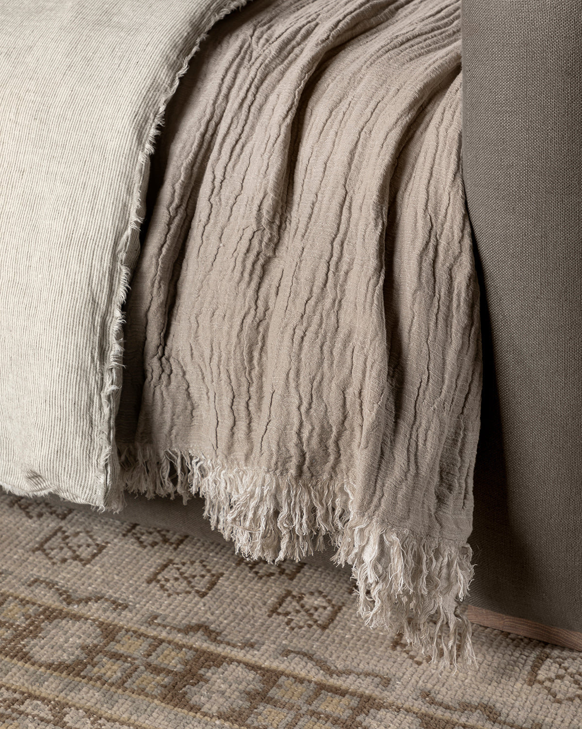 A close-up of the Ermessa Oversized Throw, a crinkled beige and off-white cotton-bamboo blend blanket, draped over a taupe upholstered bed frame beside a patterned woven rug.