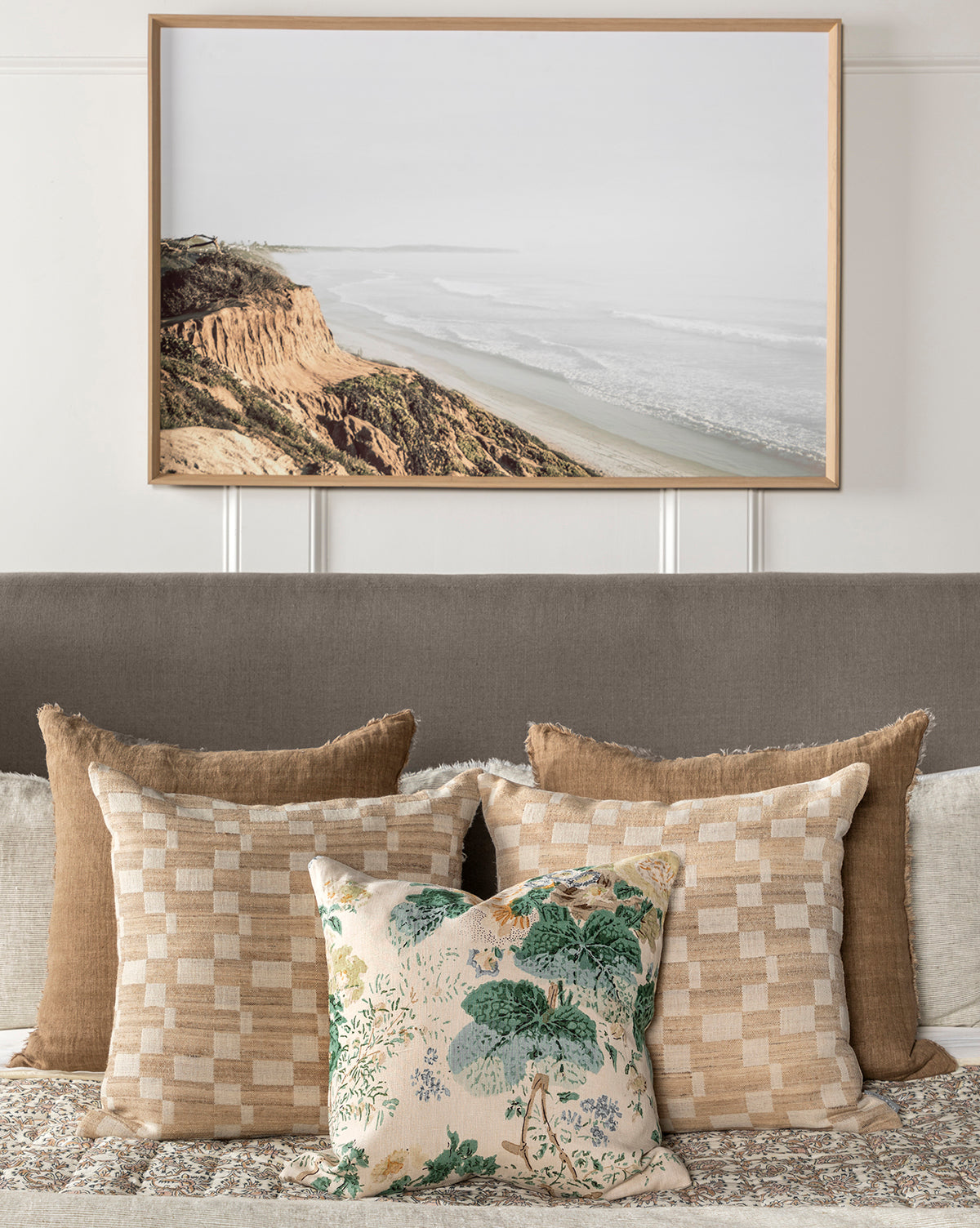The Distant Coastline framed wall art hangs above a bed adorned with beige, tan, and patterned pillows—including one with a green botanical print—complementing the room’s earthy, neutral decor palette.