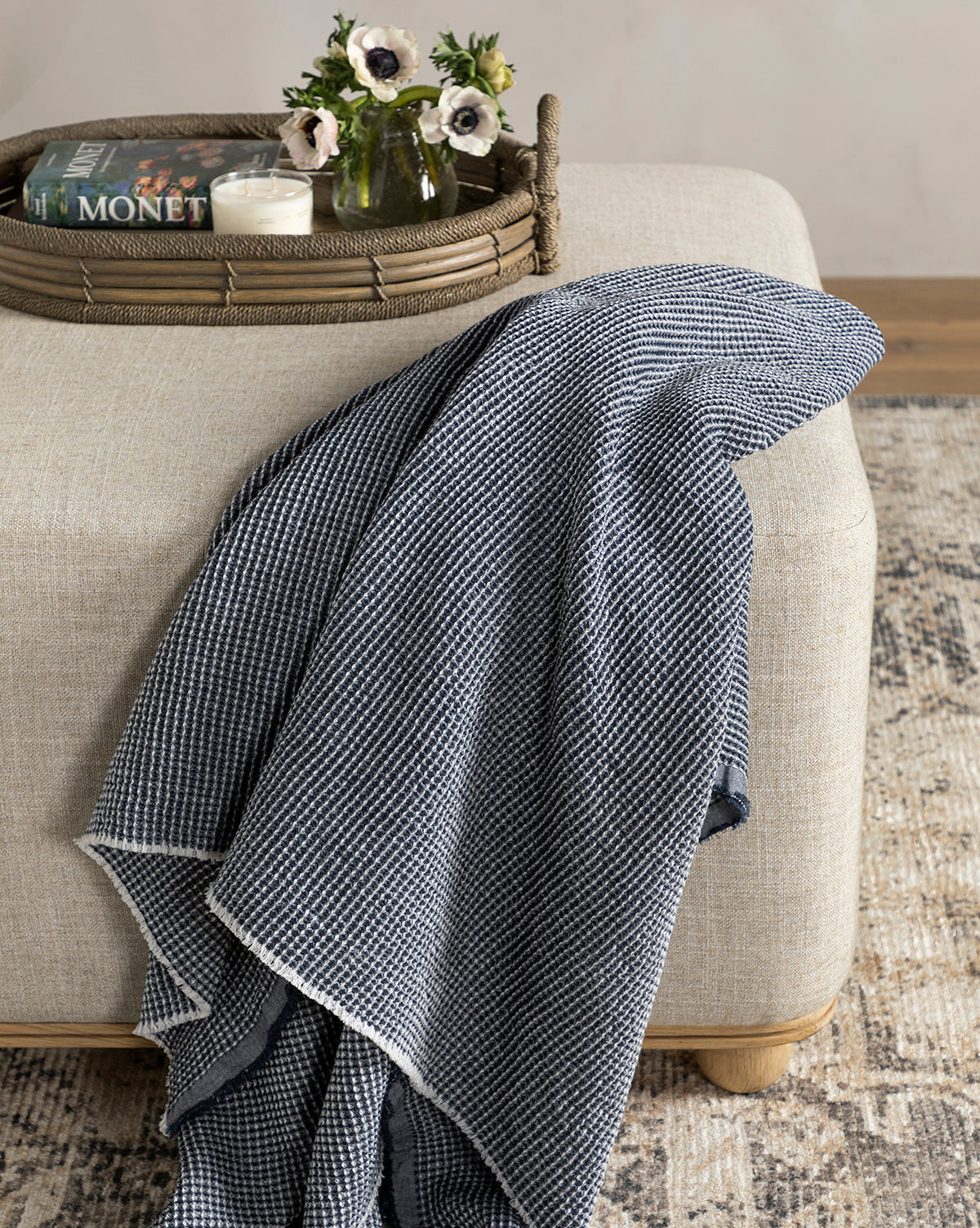 The Rebecca Throw, a blue and white cotton piece with waffle weave texture, is draped over a light beige ottoman. A tray on top displays a candle, the book Monet, and a small vase of white flowers.