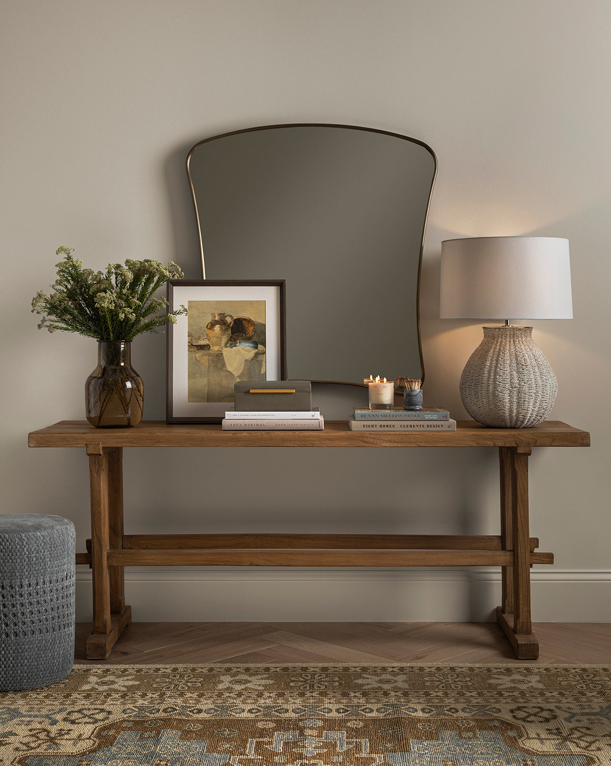 A wooden console table topped with a vase, art, books, candles, and a textured lamp sits against a beige wall with the Ludwig Wall Mirror in brass above it; nearby are a patterned rug and pouf by McGee & Co.