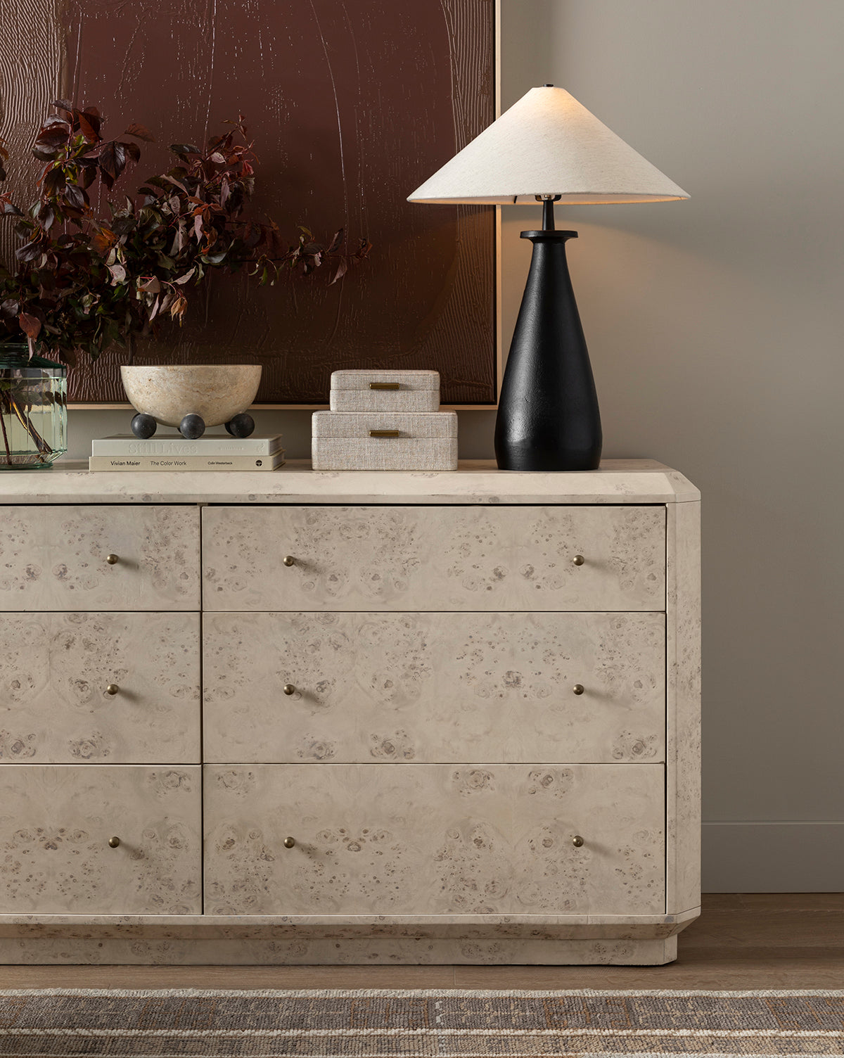 A beige dresser with brass knobs holds the Innes Tapered Table Lamp, two decorative boxes, a bowl, a book, and a vase with dark foliage. Above, a rectangle mirror on the taupe wall reflects abstract art behind by McGee & Co.